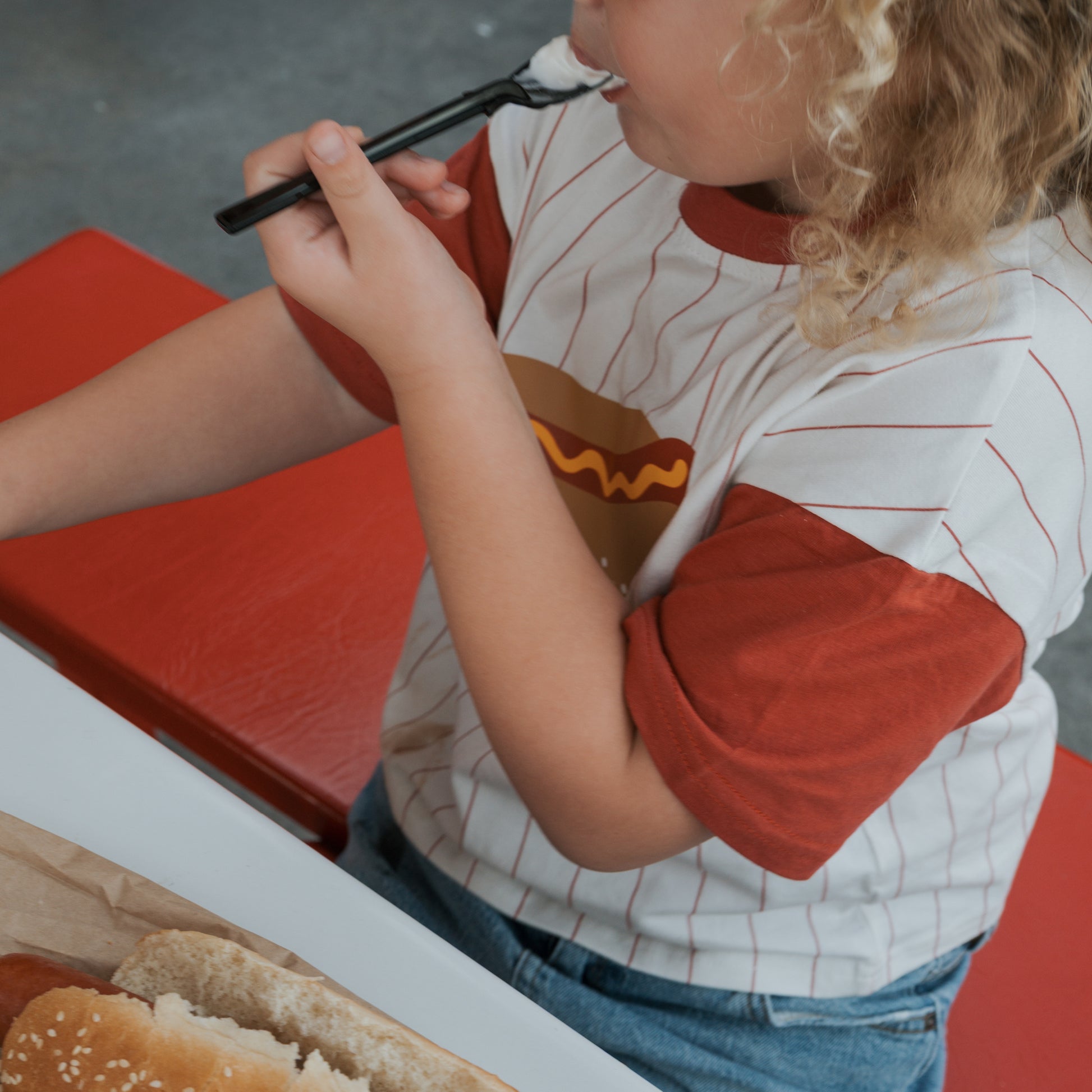 A young child in a forever french baby Child Cotton Tee | Hot Dogs sits at a red table eating with a spoon, with a hot dog bun on brown paper beside them.