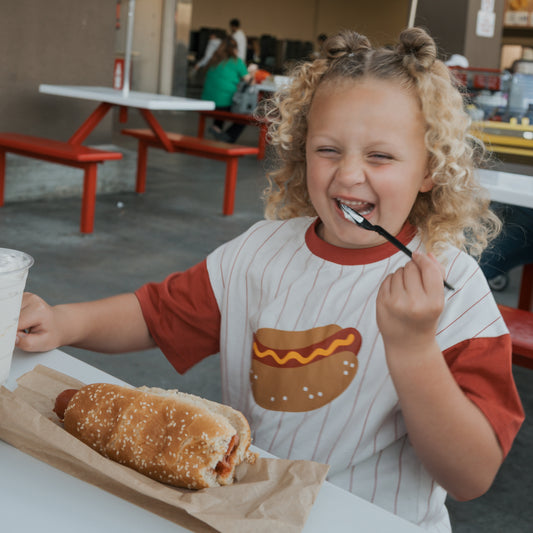 A happy child in the forever french baby Child Cotton Tee | Hot Dogs, featuring a playful cartoon hot dog, sits at a table ready to enjoy a hot dog and drink—making mealtime extra fun in their matching kids outfit.