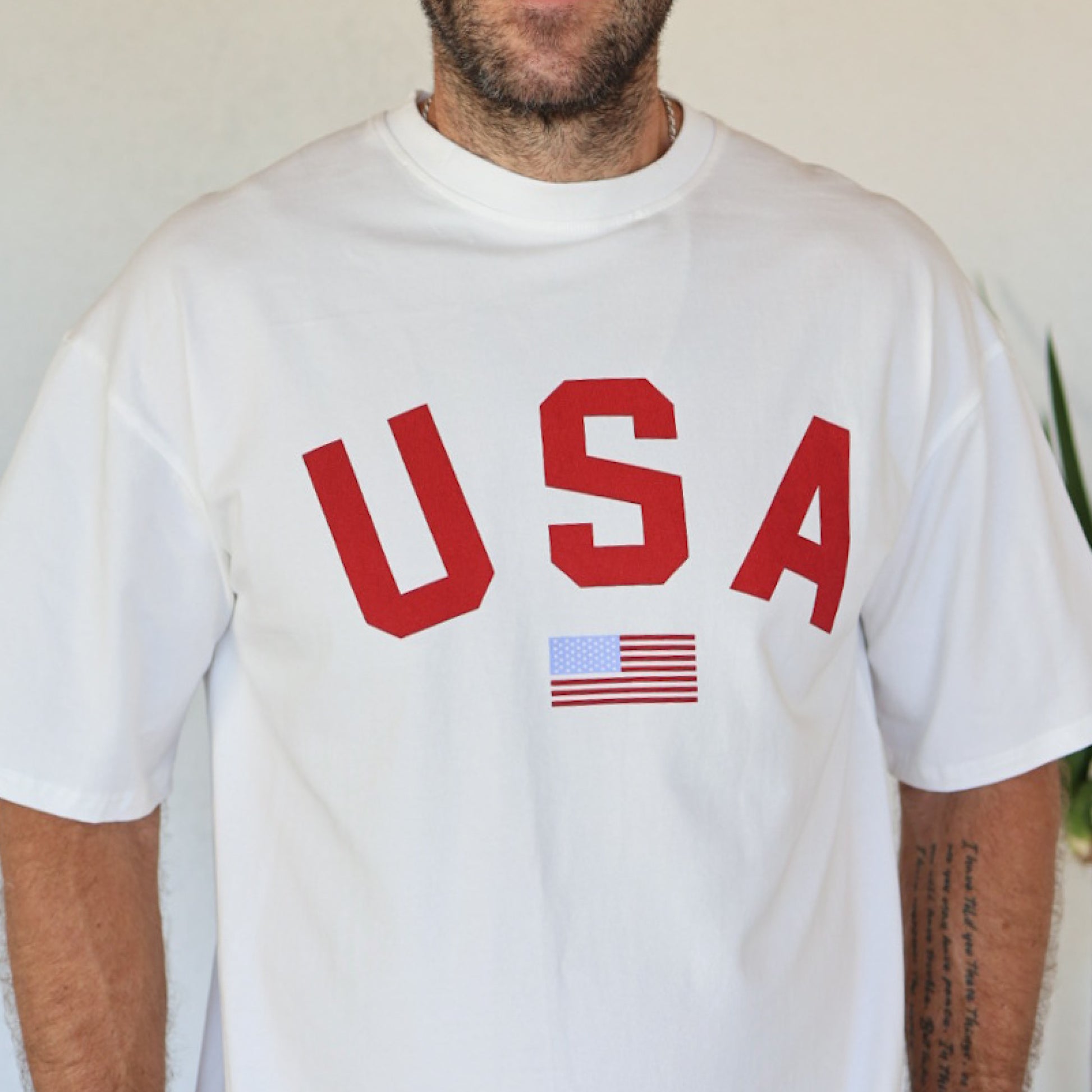 A person wears the "Adult Cotton Tee | USA" by forever french baby, featuring bold red "USA" lettering above a small American flag; their face is partially out of frame, revealing tattoos on their right forearm.