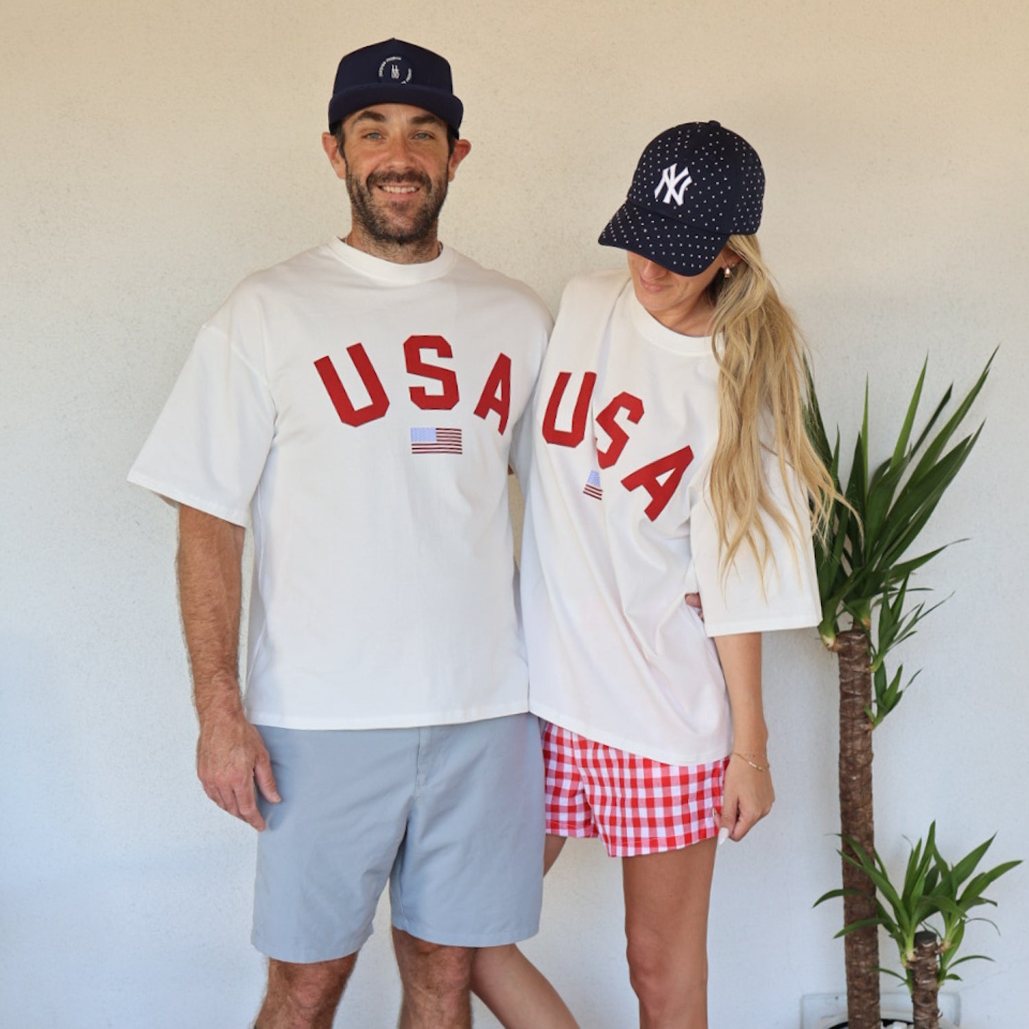 A man and woman wear matching forever french baby Adult Cotton Tee | USA shirts. He pairs his 100% cotton tee with shorts and a navy cap; she styles hers with red checkered shorts and a Yankees cap, showing off this classic unisex look.