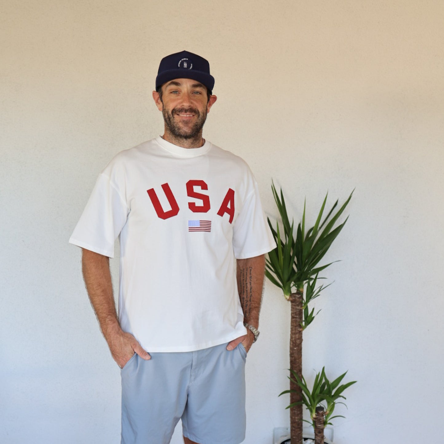 A smiling man in a blue hat and gray shorts stands indoors by a green plant, wearing the forever french baby Adult Cotton Tee | USA—an all-cotton unisex shirt with "USA" and an American flag—against a plain light wall.