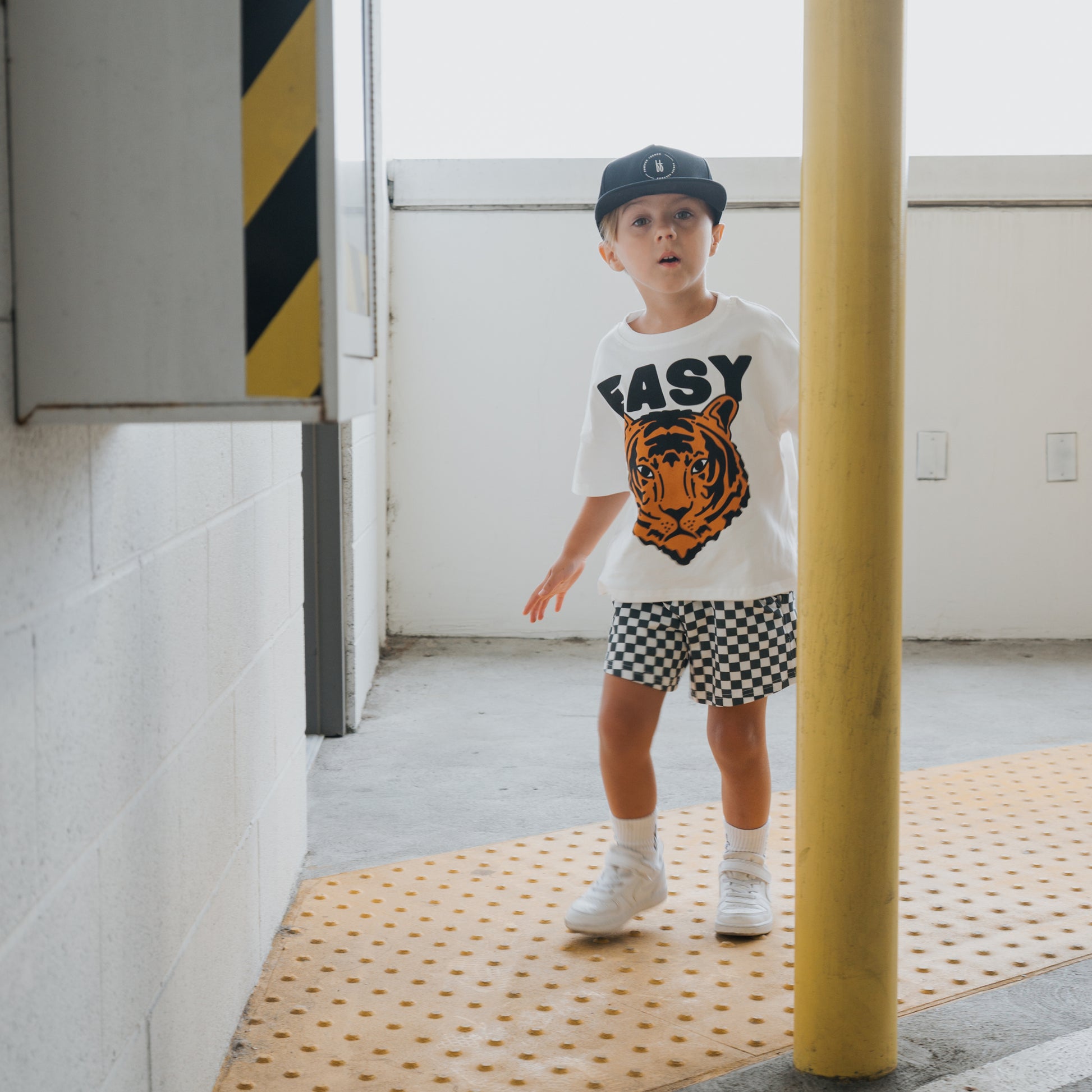 A young child in the forever french baby Child Short Set | Easy Tiger—featuring a tiger graphic tee marked "EASY" and checkered shorts—stands indoors by a yellow pole and tactile paving, wearing a black cap and white sneakers.