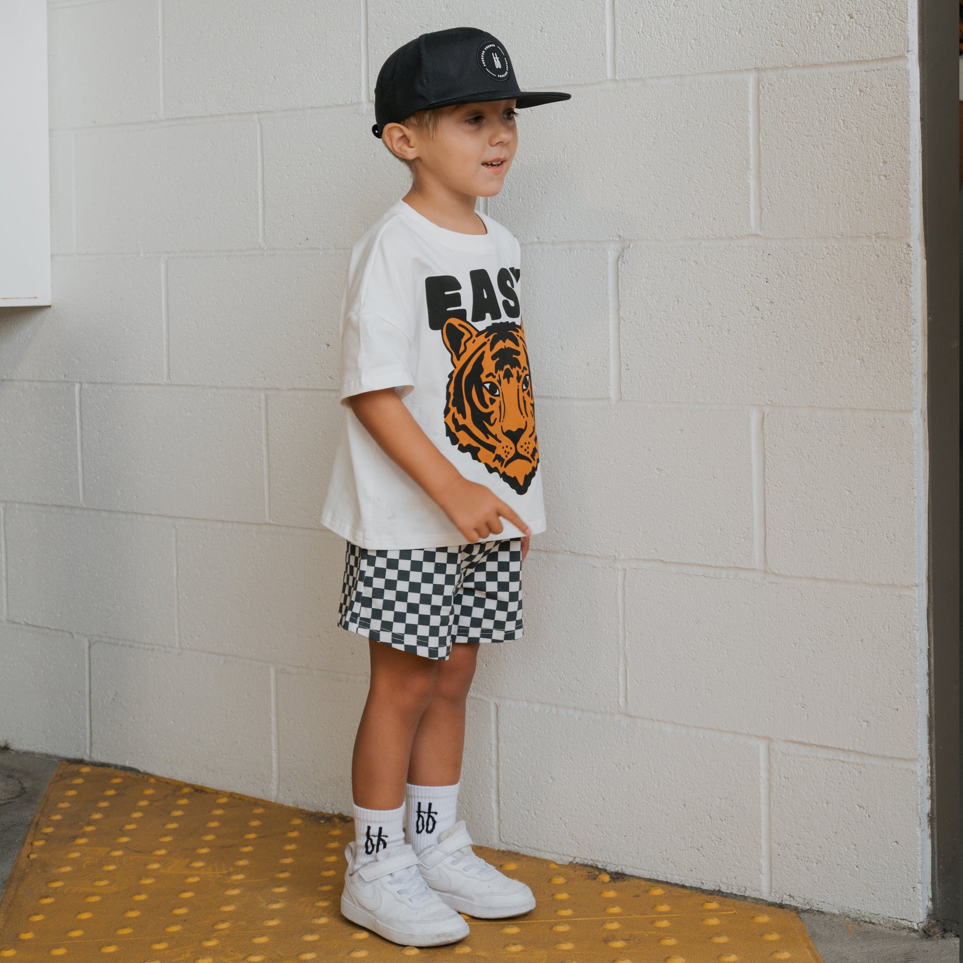 A young boy stands on a yellow mat by a white brick wall, wearing the forever french baby Child Short Set | Easy Tiger and accessorizing with a black cap, white "bb" socks, checkered shorts, and white sneakers.