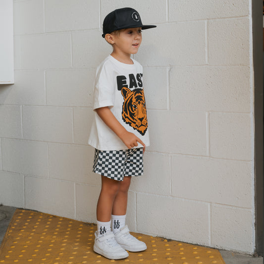 A young boy stands on a yellow mat by a white brick wall, wearing the forever french baby Child Short Set | Easy Tiger and accessorizing with a black cap, white "bb" socks, checkered shorts, and white sneakers.