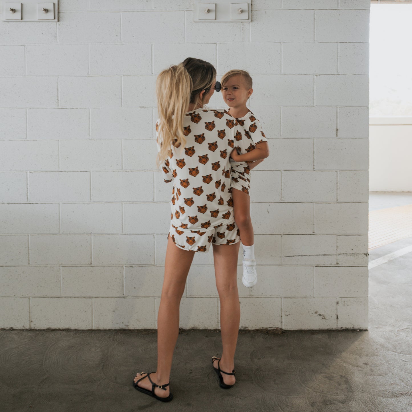 A woman wearing forever french baby Women's Bamboo Short Set in Tigers print holds a child in matching breathable pajamas, both facing away from the camera in front of a white cinder block wall.