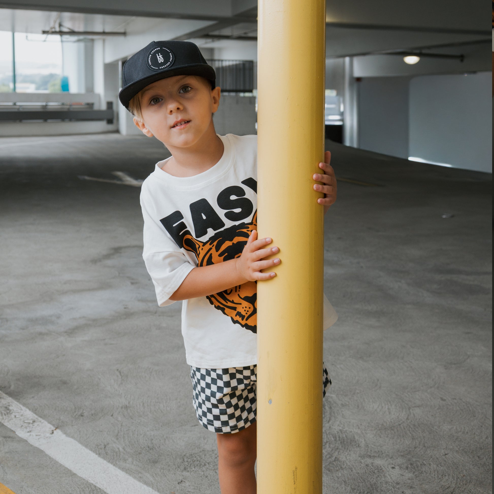 A young boy in the forever french baby Child Short Set | Easy Tiger—featuring a tiger graphic shirt and checkered shorts—stands in a parking garage, playfully peeking from behind a yellow pole.