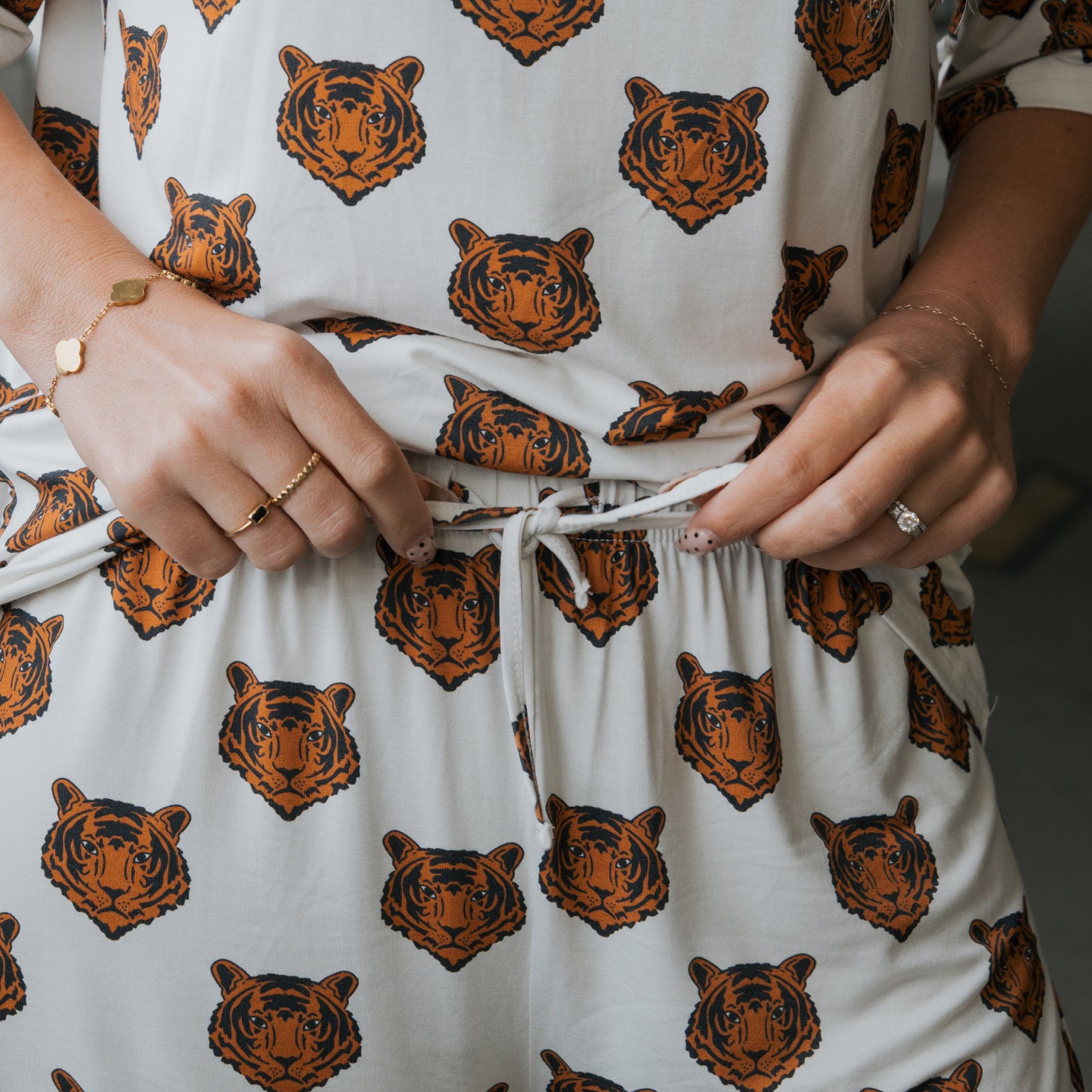 A person ties the drawstring of the Women's Bamboo Short Set | Tigers by forever french baby, featuring white, breathable bamboo fabric with orange and black tiger faces. They accessorize with rings and bracelets.