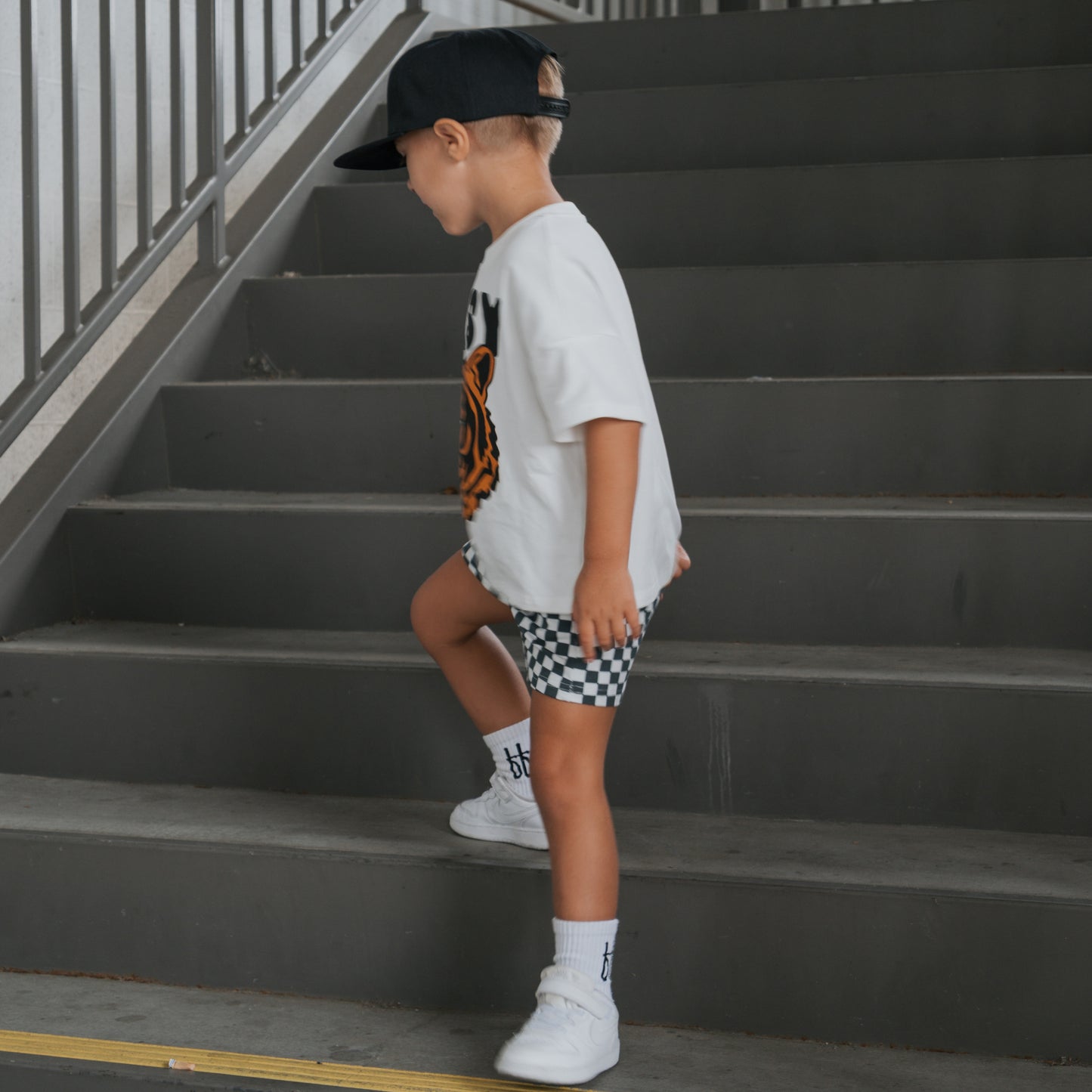 A child in a forever french baby Child Short Set | Easy Tiger—a cotton blend white t-shirt with an orange graphic and shorts—walks up gray stairs in white sneakers, socks, and a black cap beside a metal railing.