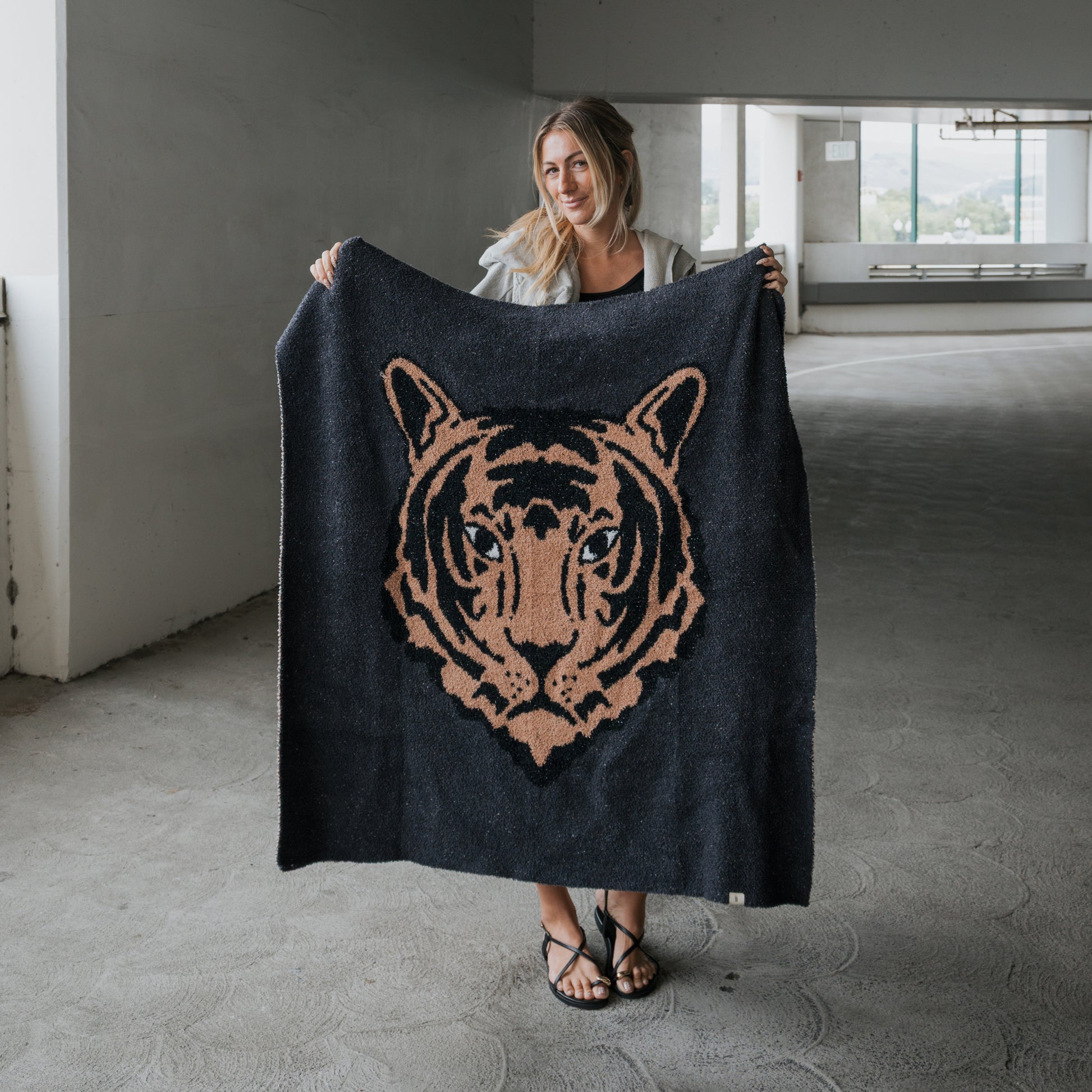 A woman in a parking garage holds up the forever french baby Plush Blanket | Tiger, a soft microfiber throw with a large stylized tiger face design in tan and black.