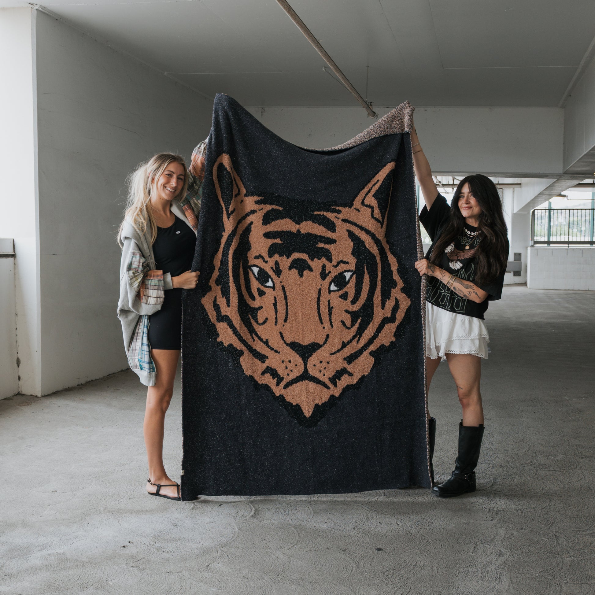 Two women in casual attire stand in a parking garage, smiling as they display the forever french baby Plush Blanket | Tiger, which showcases a detailed tiger face illustration and features soft microfiber material.