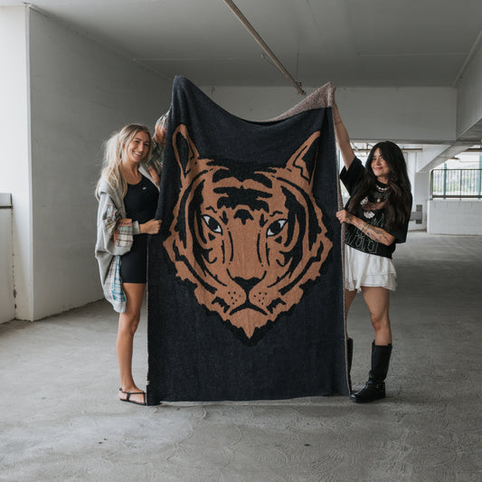 Two women in casual attire stand in a parking garage, smiling as they display the forever french baby Plush Blanket | Tiger, which showcases a detailed tiger face illustration and features soft microfiber material.