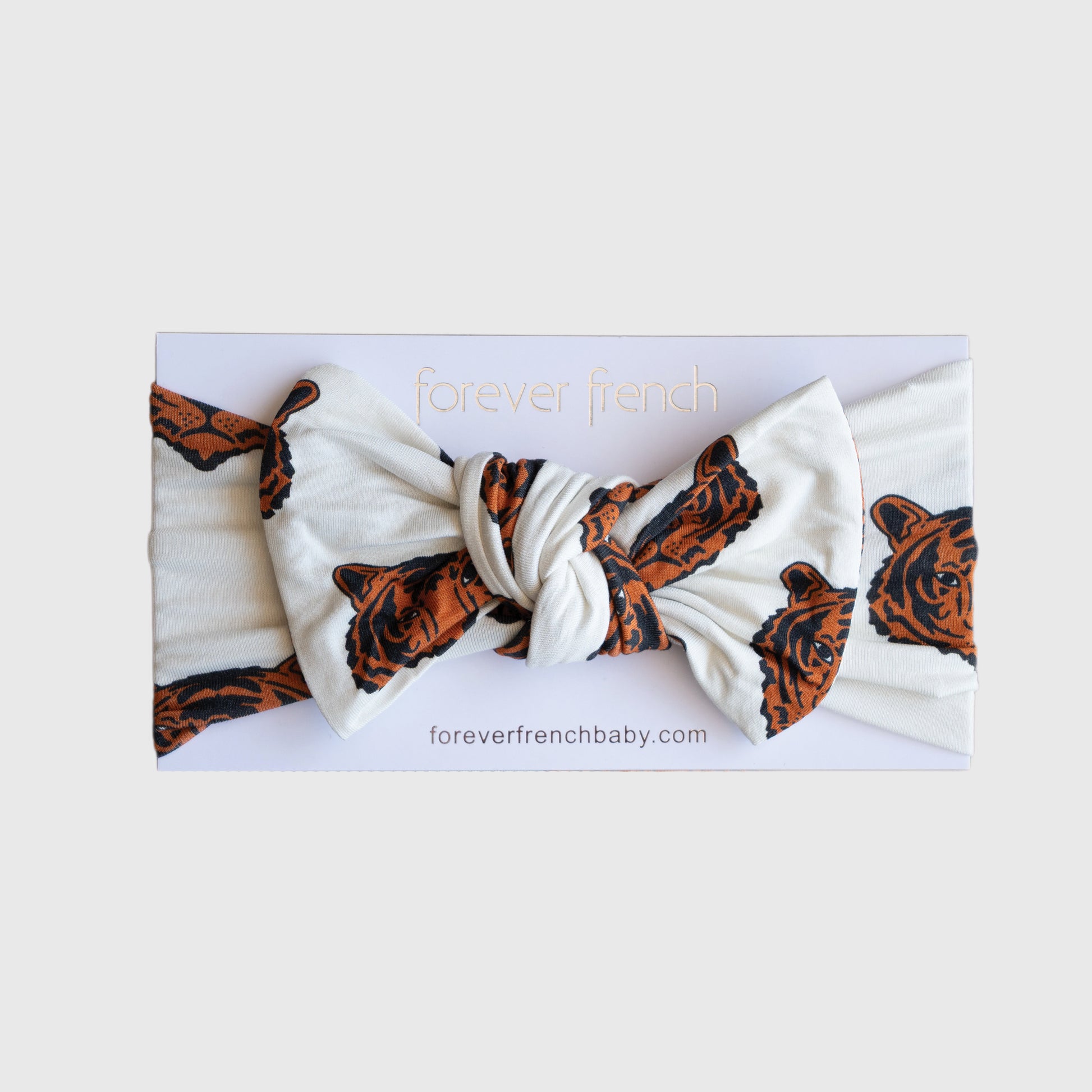 The Bamboo Head Wrap | Tigers by forever french baby features a white adjustable band with brown tiger faces tied in a large bow, displayed on branded packaging with "foreverfrenchbaby.com" at the bottom.