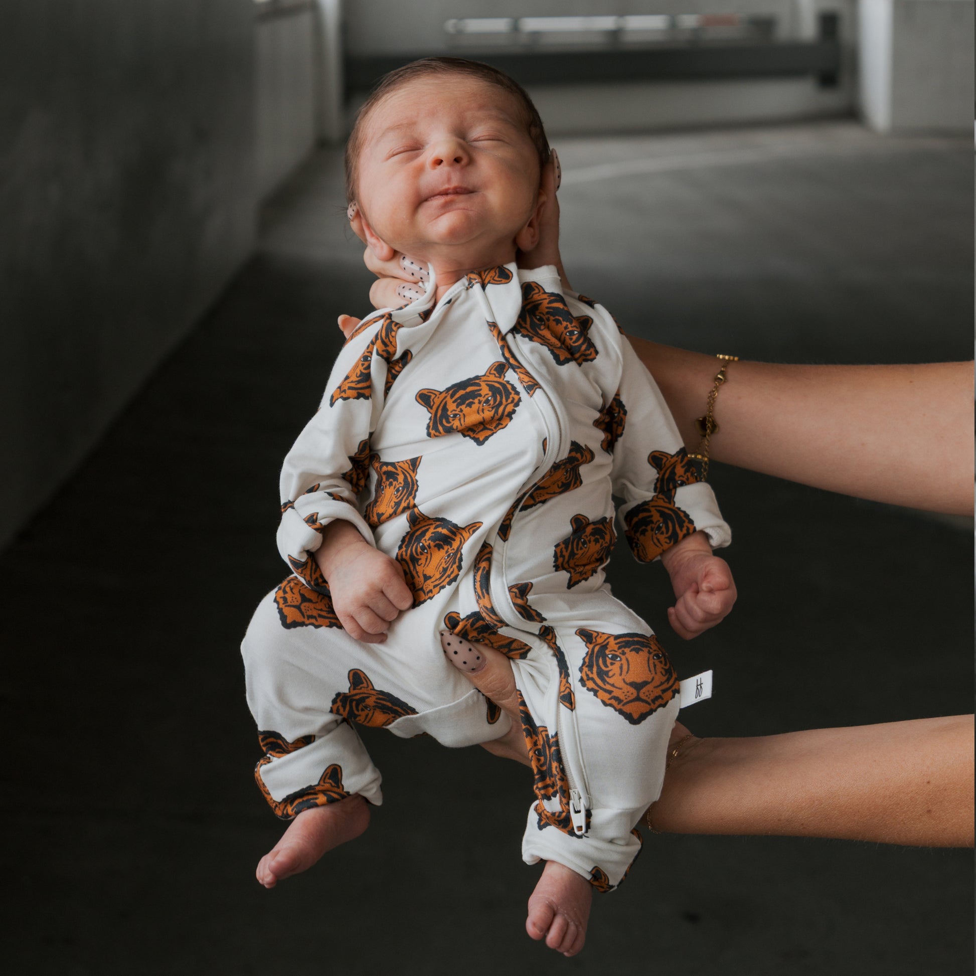 A newborn sleeps peacefully in forever french baby Bamboo Zip Pajamas | Tigers, featuring soft, breathable fabric with tiger faces, gently held by an adult in a dimly lit room.