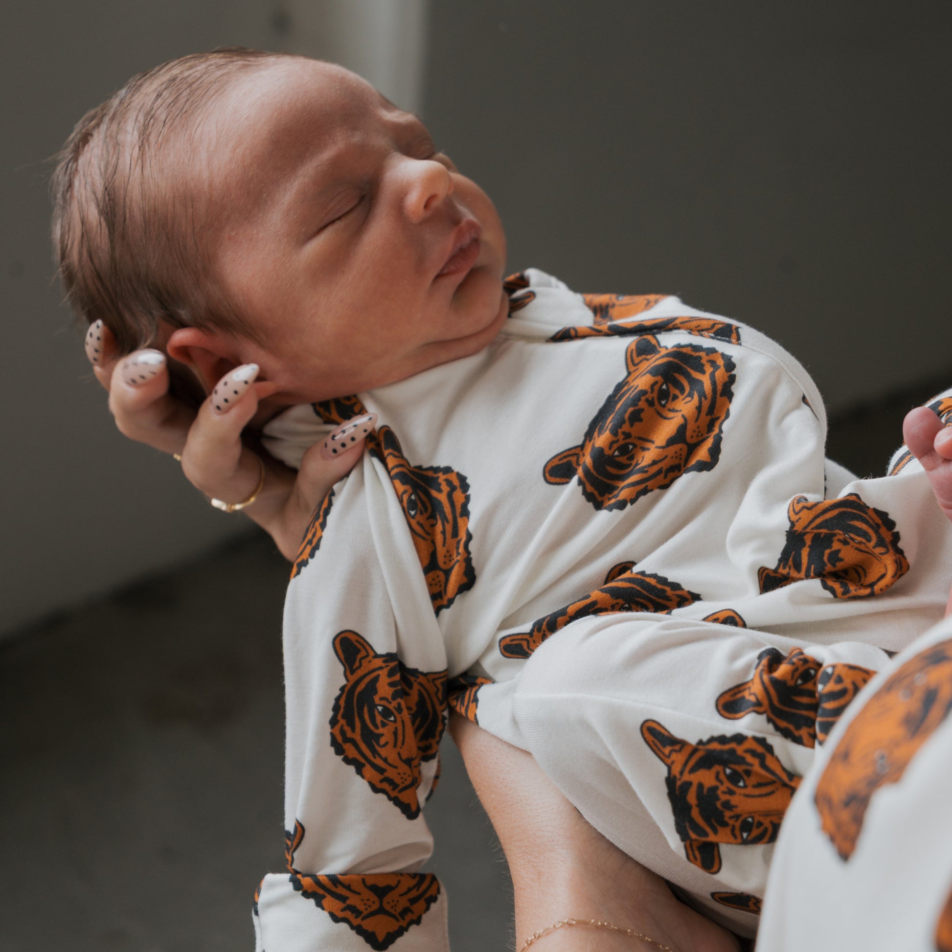 A newborn sleeps peacefully in forever french baby Bamboo Zip Pajamas | Tigers, featuring orange and black tiger faces, while being gently held by an adult with painted nails.