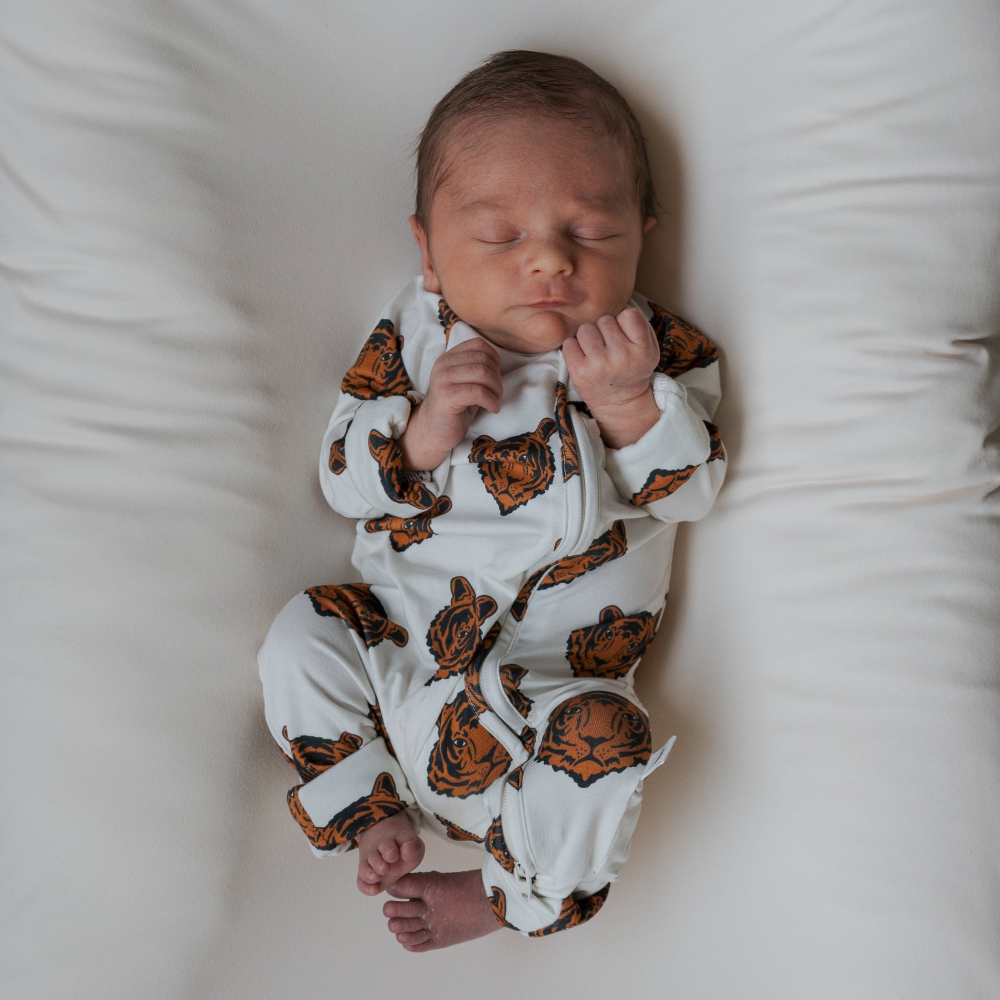 A newborn sleeps on a soft white surface, dressed in forever french baby Bamboo Zip Pajamas | Tigers—a white onesie with orange tiger faces. The baby's hands rest near its face and its legs are relaxed.