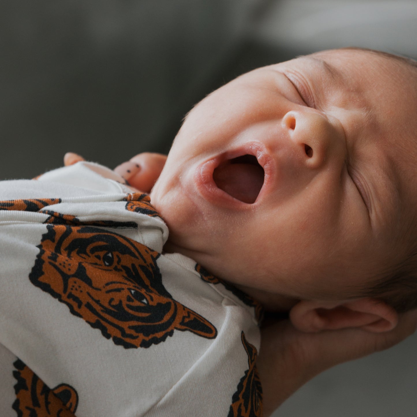 A close-up of a yawning baby in forever french baby's Bamboo Zip Pajamas | Tigers, featuring a tiger face print, being gently held by someone’s hand.