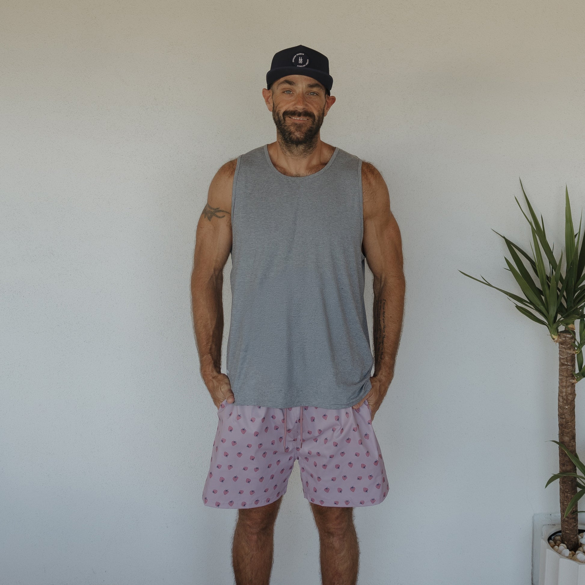 A smiling man in a navy cap, gray sleeveless shirt, and forever french baby Men's Board Short | Matilda stands against a white wall next to a potted plant.