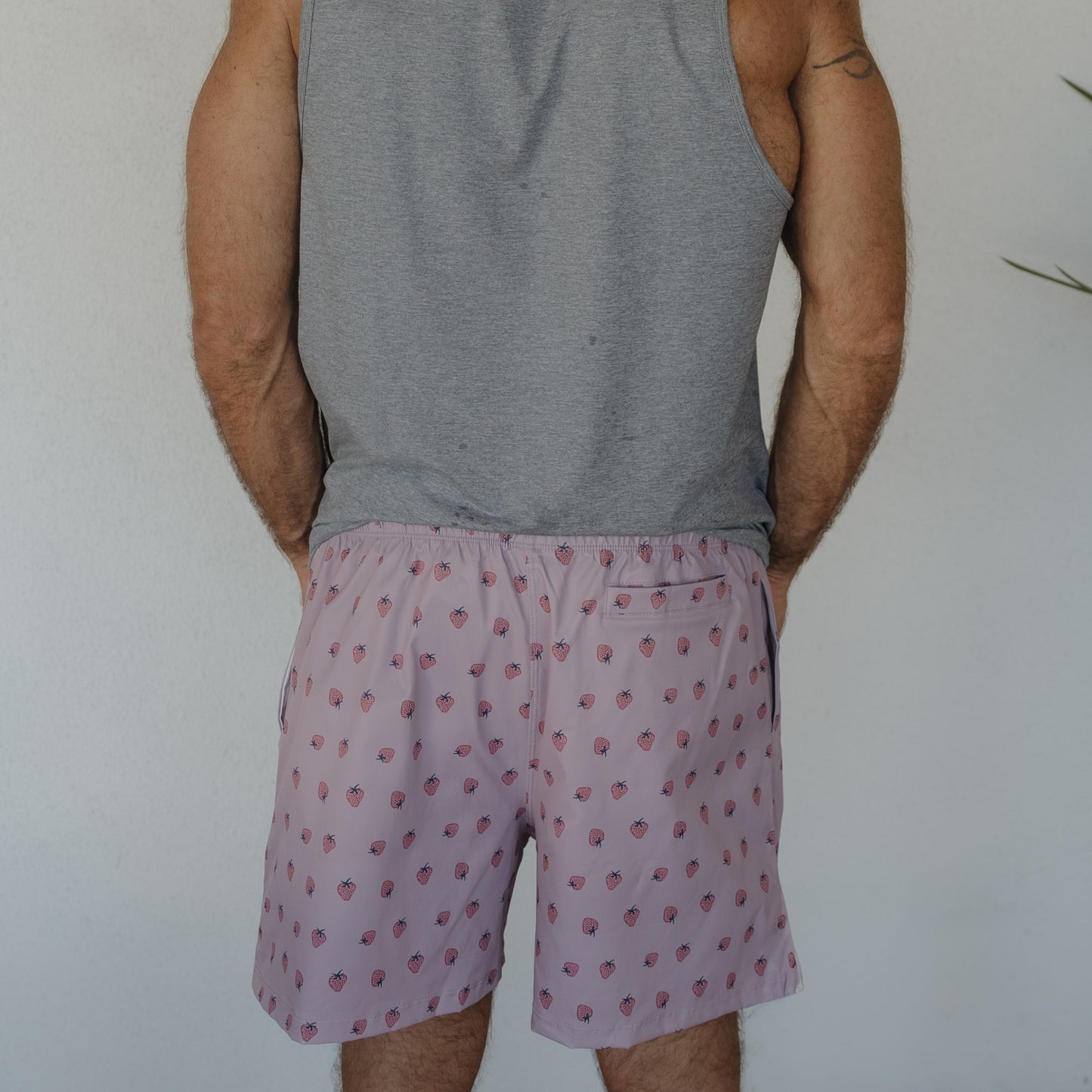 A person in a gray sleeveless shirt and forever french baby Men's Board Short | Matilda stands facing away from the camera against a plain light wall.