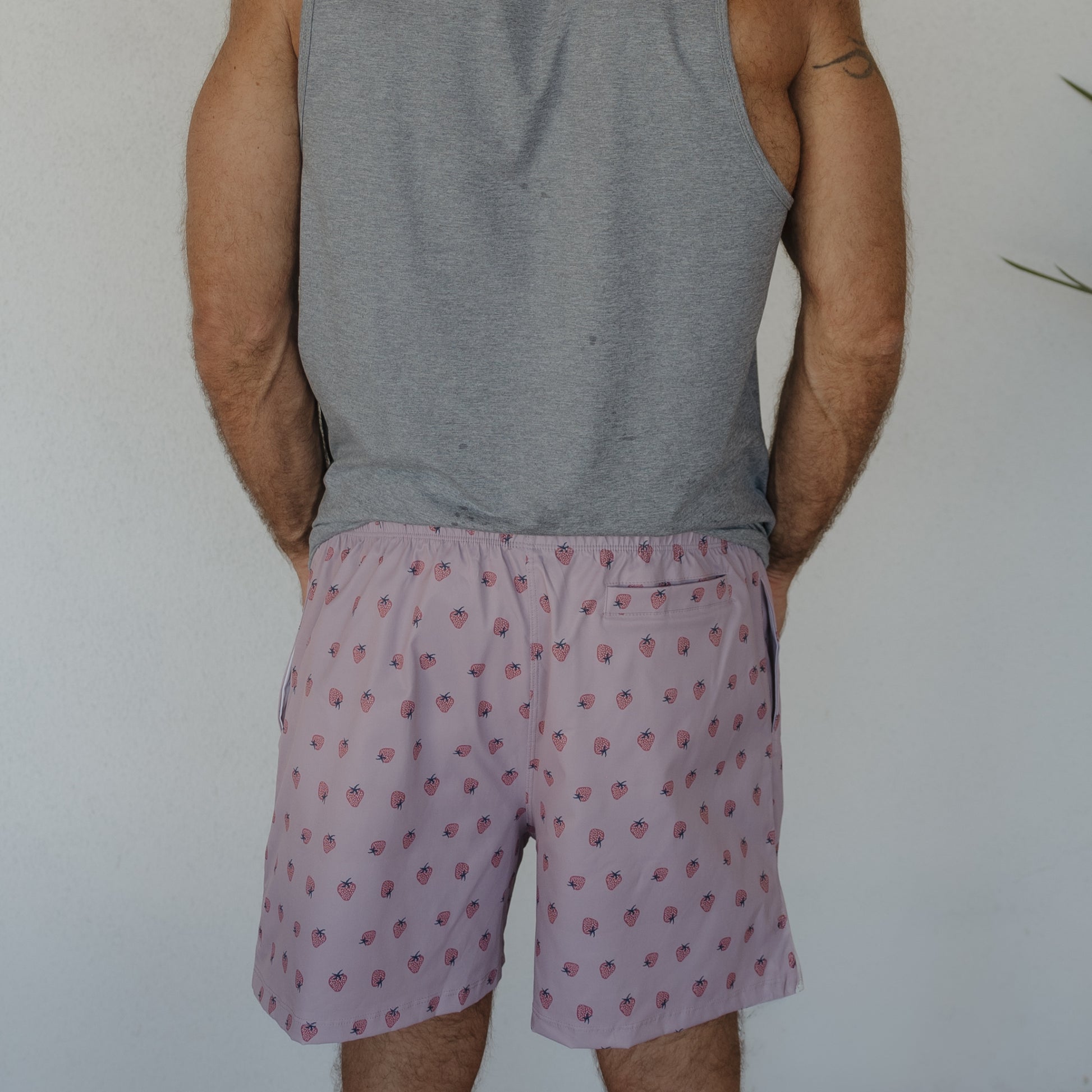 A person in a gray sleeveless shirt and forever french baby Men's Board Short | Matilda stands facing away from the camera against a plain light wall.