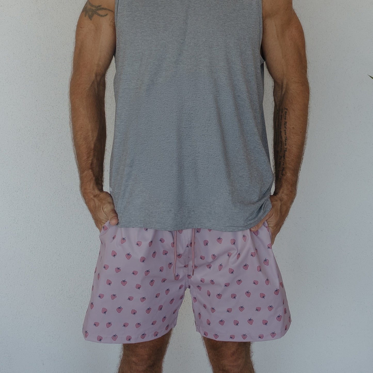 A man models the Men's Board Short | Matilda by forever french baby, featuring a pink strawberry print and gray sleeveless shirt, standing against a plain white wall with his hands in his pockets. His face is not visible.