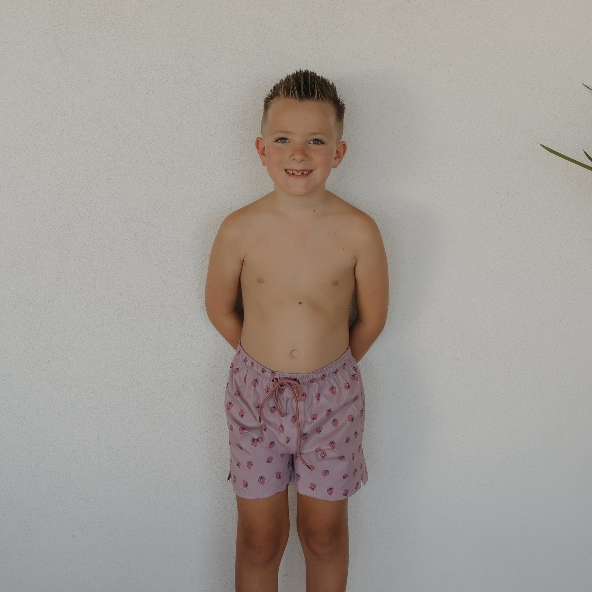 A young boy with short hair stands barefoot against a white wall, smiling and wearing forever french baby's Child Board Short | Matilda—light pink with a subtle pattern and perfect for kids swimwear. A green plant appears on the right edge.