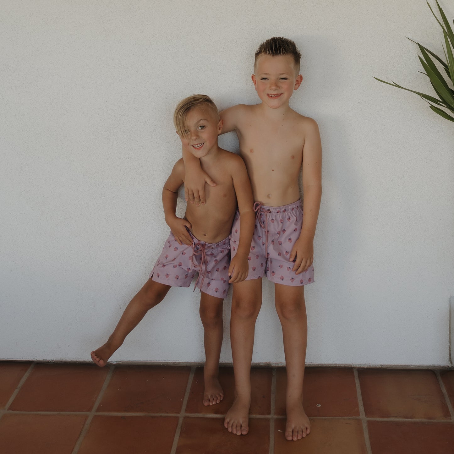 Two young boys in forever french baby’s Child Board Short | Matilda, featuring a mauve heart pattern, stand barefoot on terracotta tiles. One leans playfully on the other as they smile at the camera—a joyful kids swimwear moment.
