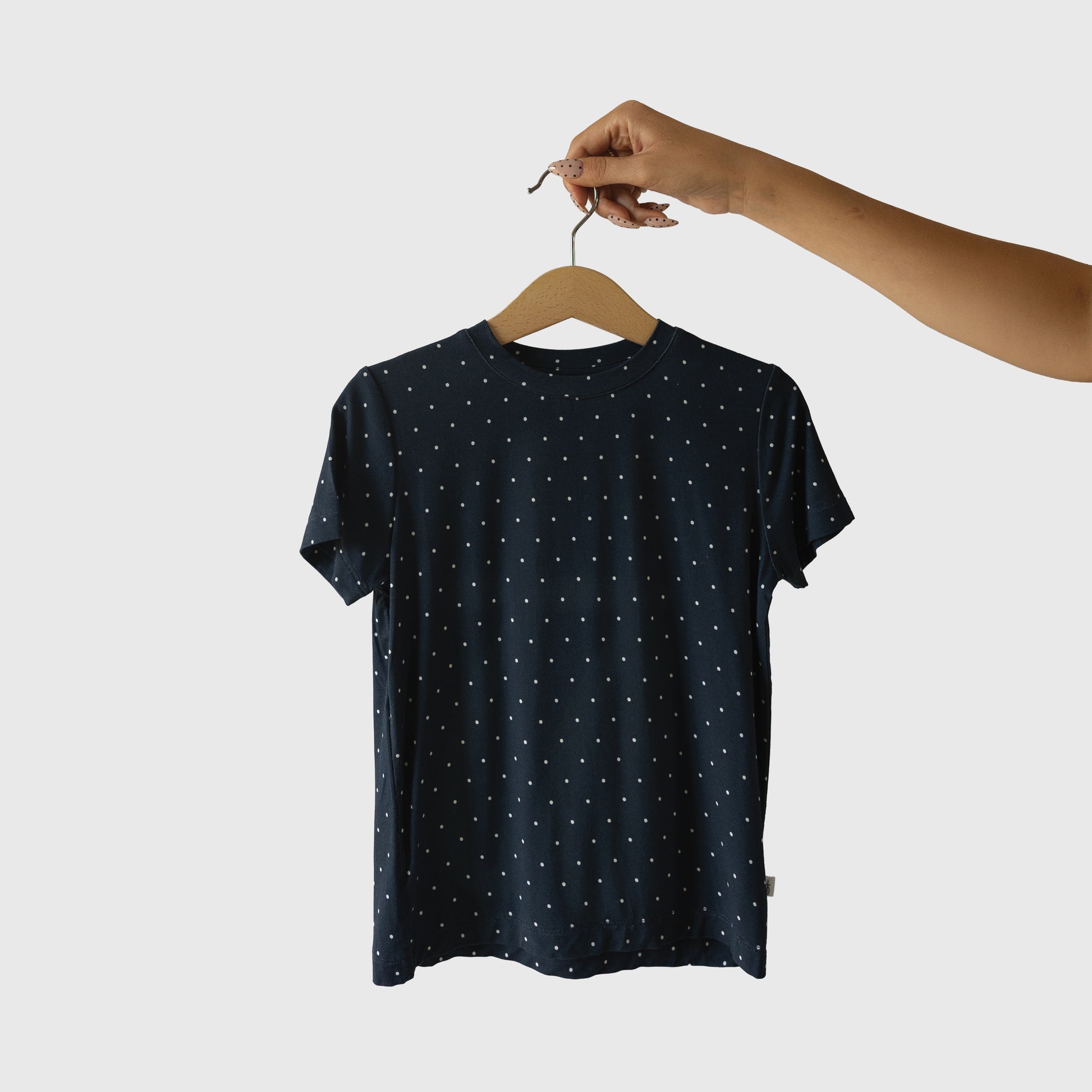 A hand holds the forever french baby Child Short Sleeve Bamboo Lounge Set | Emery, a navy blue T-shirt with small white polka dots—breathable kids' clothing—displayed against a plain white background.