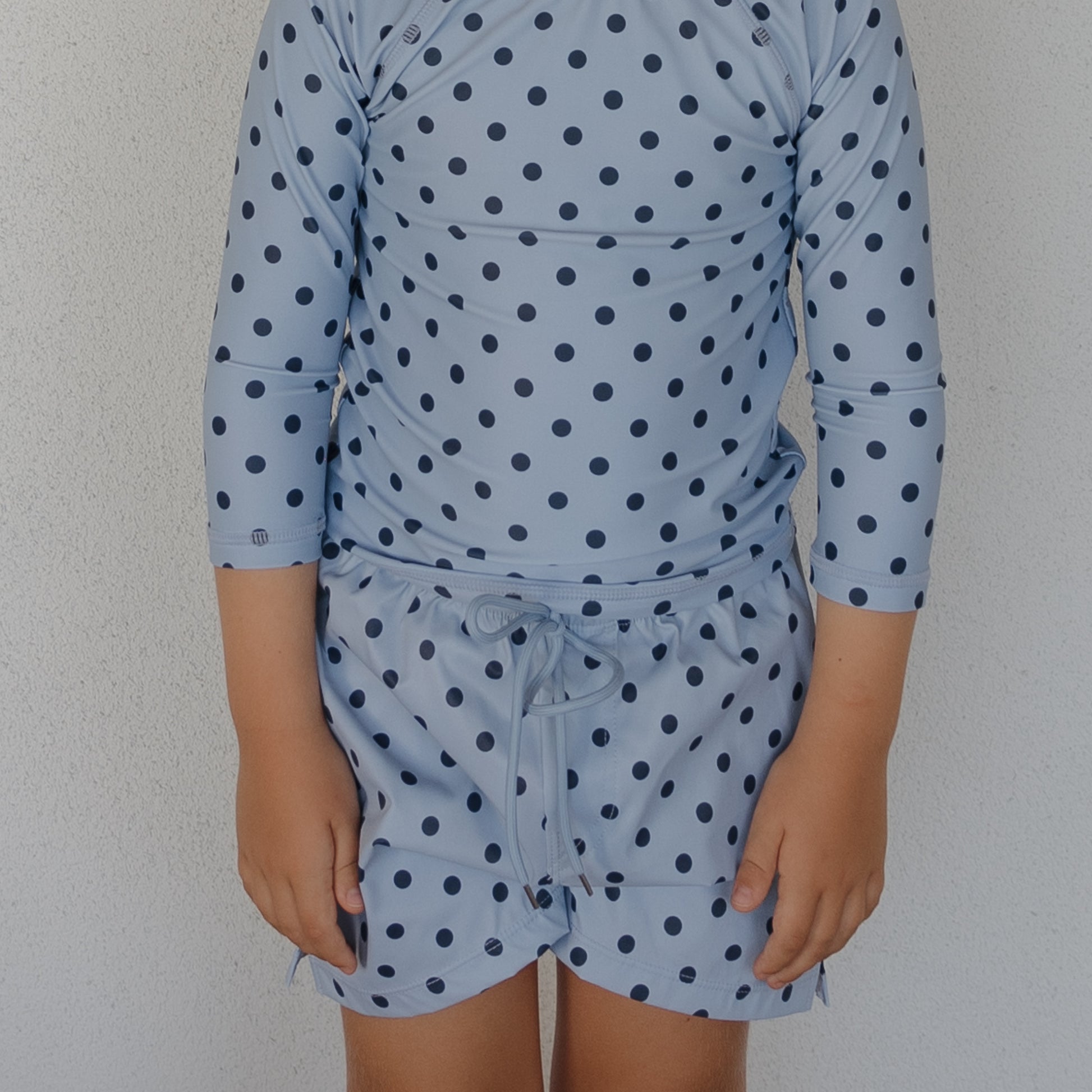 A child wearing forever french baby’s Child Board Short | Sully—a light blue, long-sleeved swim top with dark blue polka dots and matching board shorts—stands against a plain white wall. The child's face is not visible.