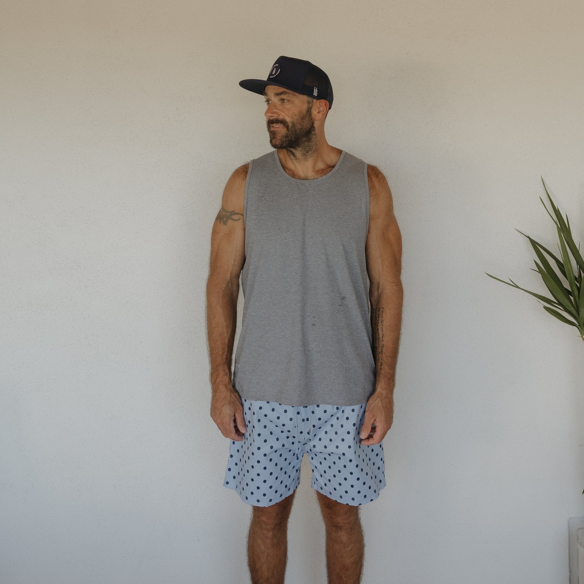 A man in a gray tank top, black cap, and forever french baby Men's Board Short | Sully stands against a white wall, glancing left. A green plant on the right hints at a stylish matching swim collection.