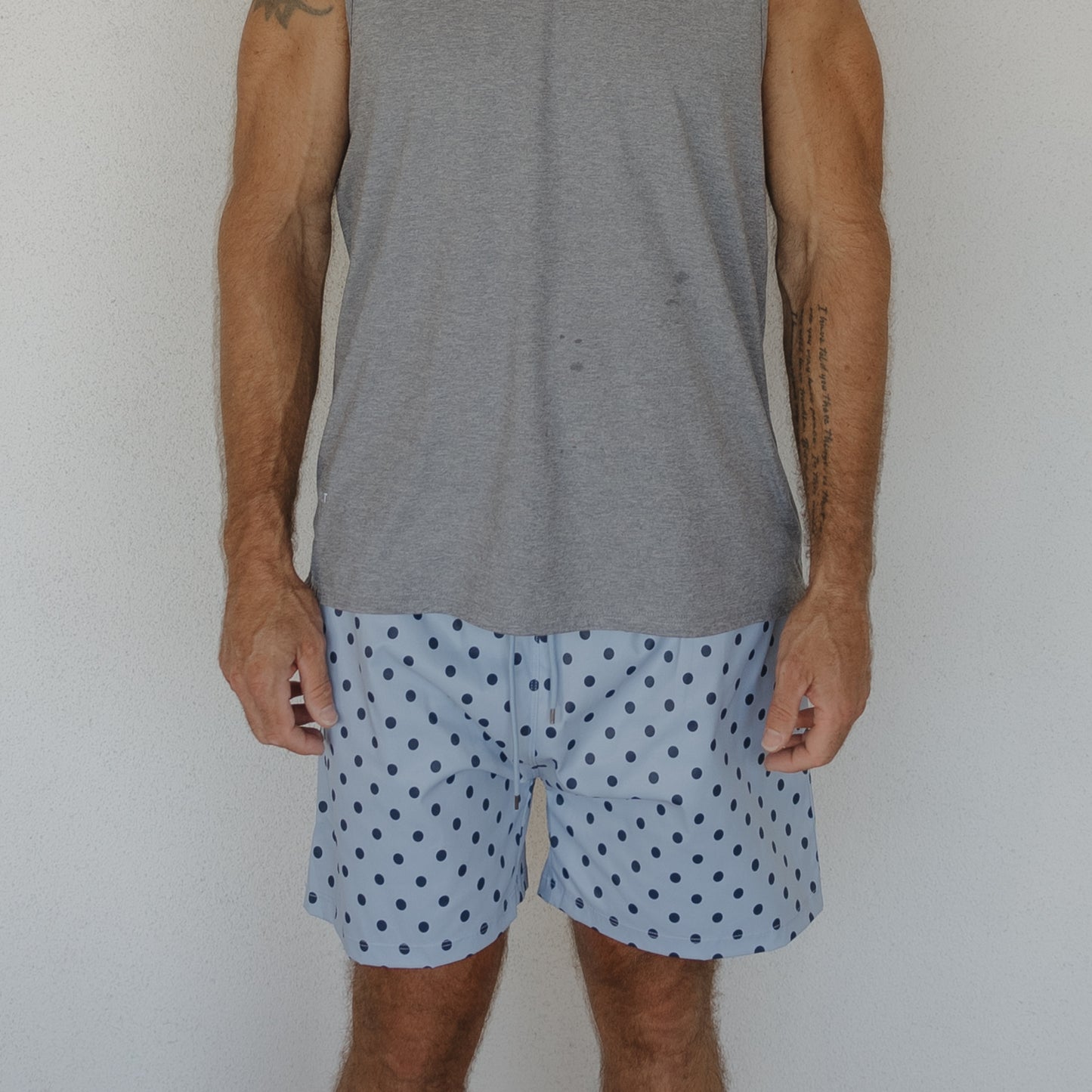 A man wearing a grey sleeveless shirt and forever french baby Men's Board Short | Sully stands against a white wall, arms relaxed at his sides. His head is not visible in the image.