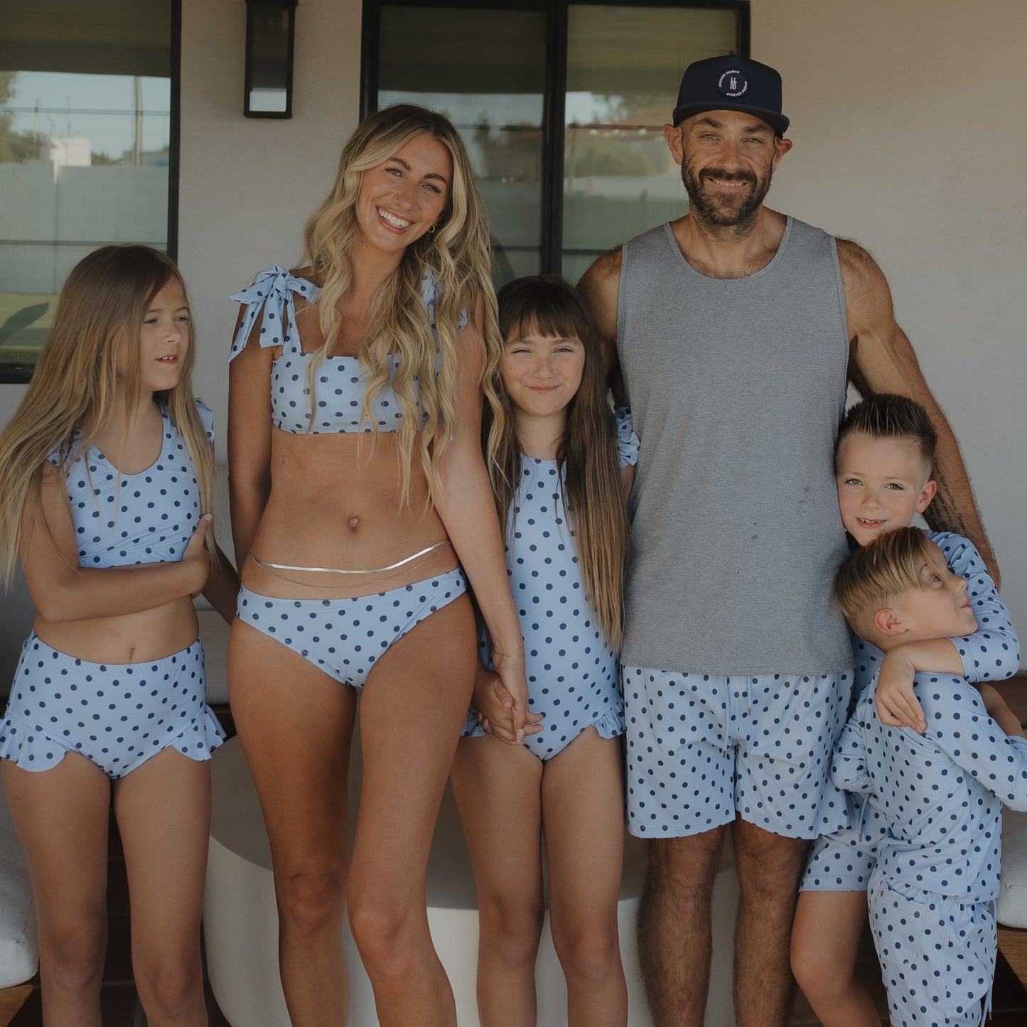A smiling family of five stands outside, happy and relaxed. The mother and three kids wear blue polka dot swimsuits, while the father and son sport coordinating Men’s Board Short | Sully from forever french baby.
