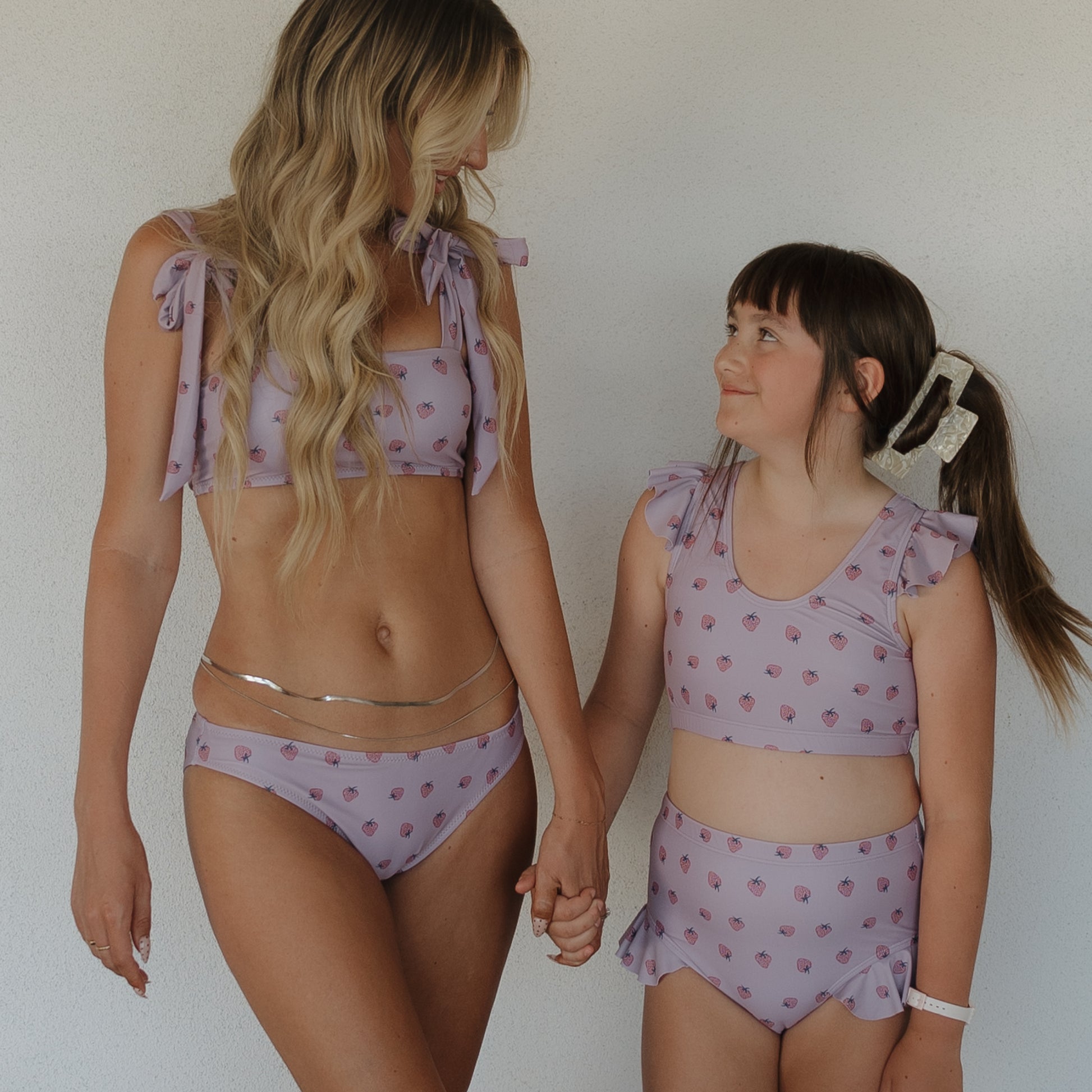 A woman and child wearing matching forever french baby Matilda two-piece swimsuits in lavender with ruffle details and a small print stand hand-in-hand, smiling at each other against a plain light background.