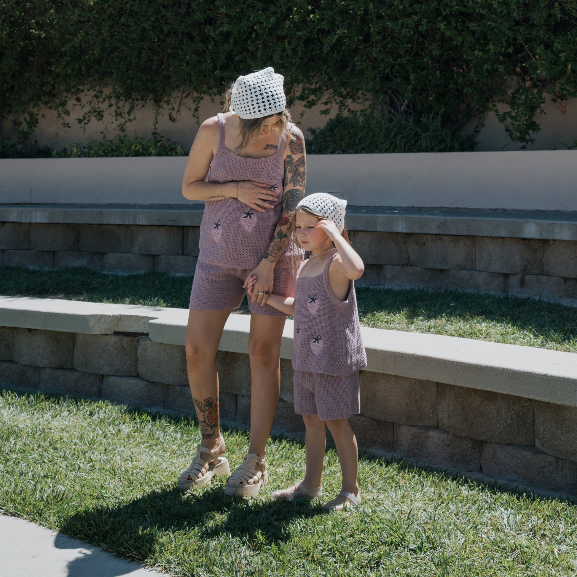 A woman wearing the forever french baby Women's Knit Short Set | Matilda in purple stands on the grass beside a child dressed to match. Both are topped with white hats; the woman cradles her baby bump while the child shields their eyes from the sun. Stone benches and lush greenery provide a scenic backdrop.