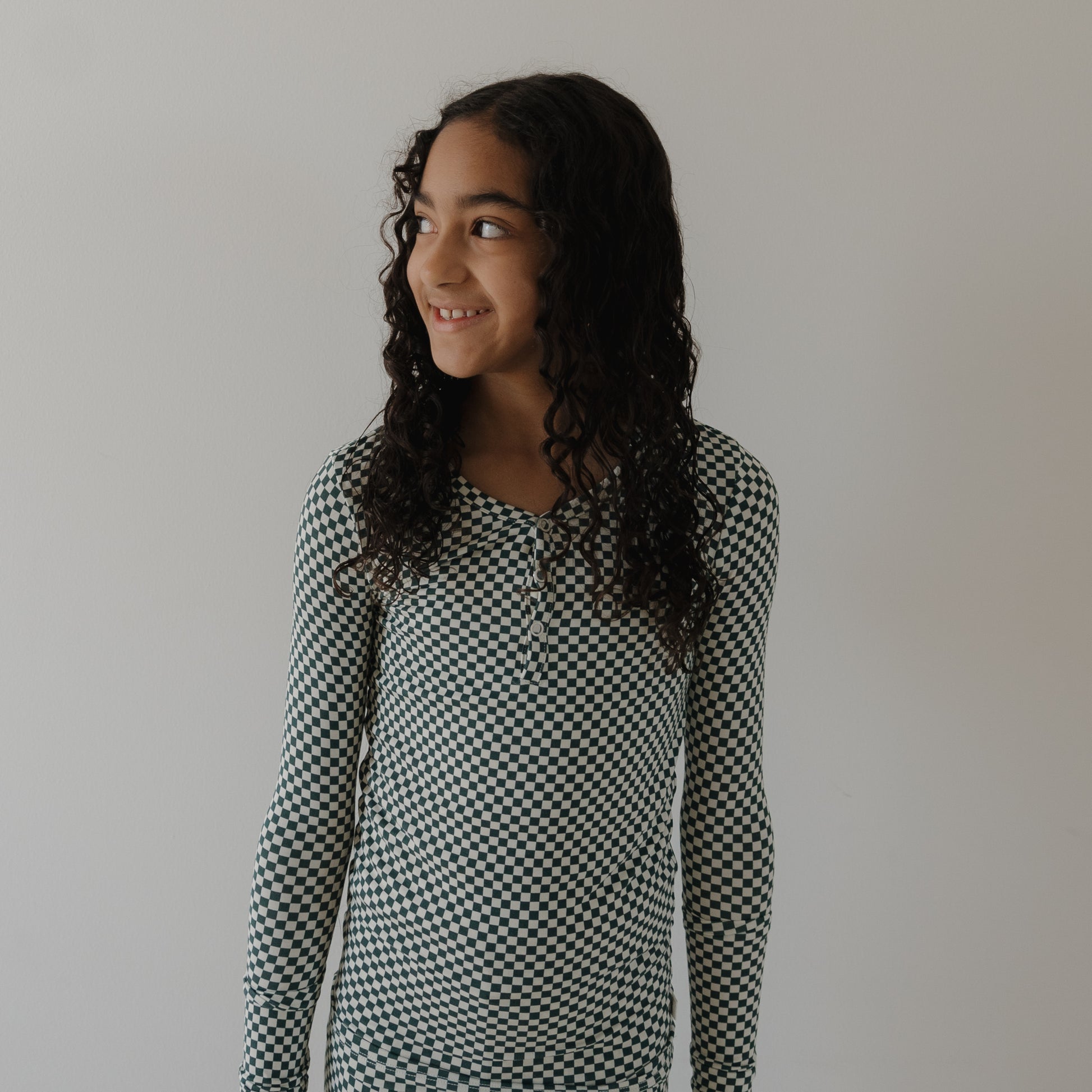 A young girl with long, curly hair wears forever french baby Bamboo Two Piece Pajamas in Lucky Micro Check and smiles to her left against a plain white wall.