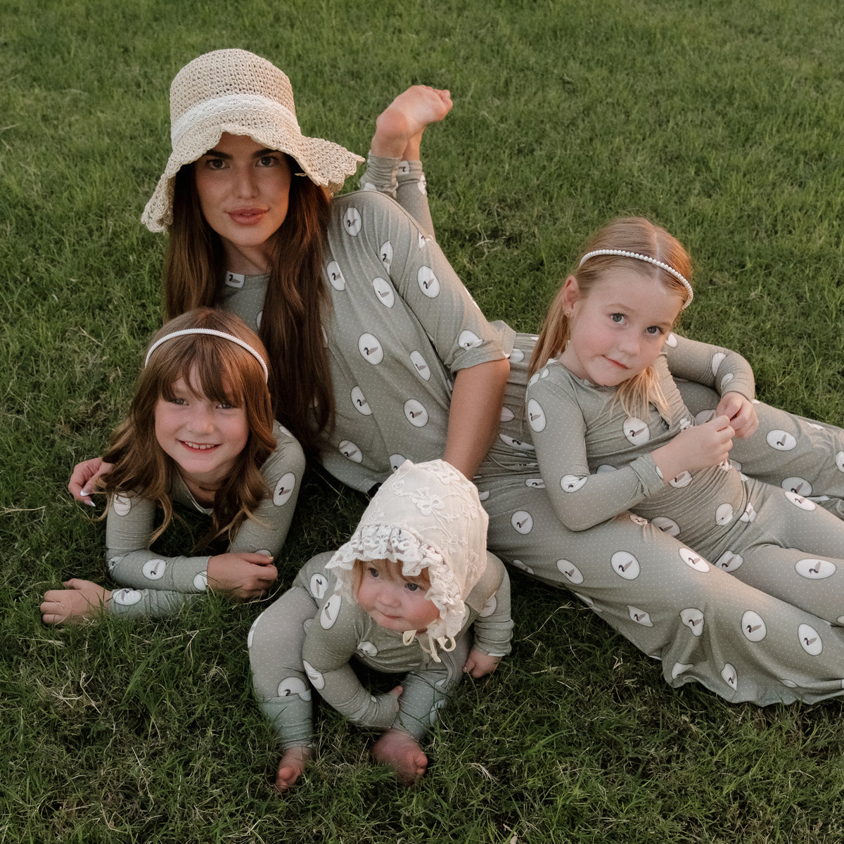 Four people, a woman and three children, all wearing matching light gray Bamboo Two Piece Pajamas | Mallard Frames from forever french baby with white polka dots, lay and sit on green grass. The woman wears a sunhat and one child wears a bonnet. Their cozy attire is crafted from hypoallergenic bamboo fabric—perfect for sensitive skin—and highlights the comfort of kids bamboo clothing by forever french baby.