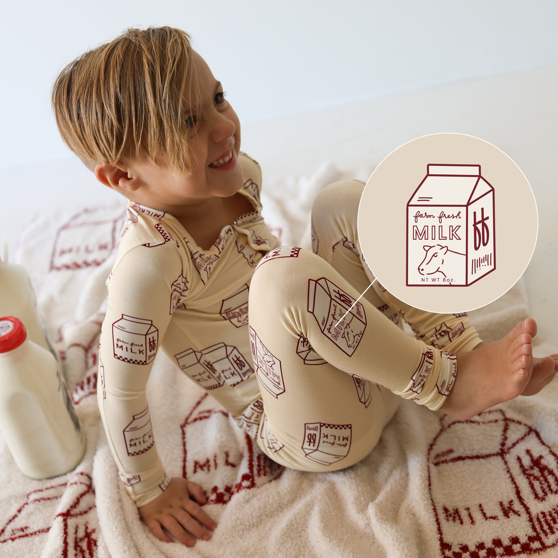 A smiling child in forever french baby’s Bamboo Two Piece Pajamas | Milk—cream bamboo PJs with red milk carton prints—sits on a milk-themed blanket beside a bottle. An inset displays the playful, hypo-allergenic design up close.