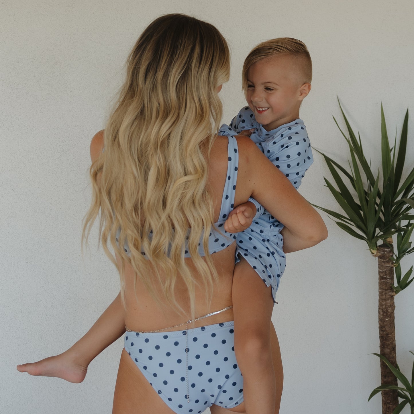 A woman with long blonde hair wears the forever french baby Women's Two Piece Swimsuit | Sully, matching her smiling child in family swimwear, as they stand by a white wall with a potted plant nearby.