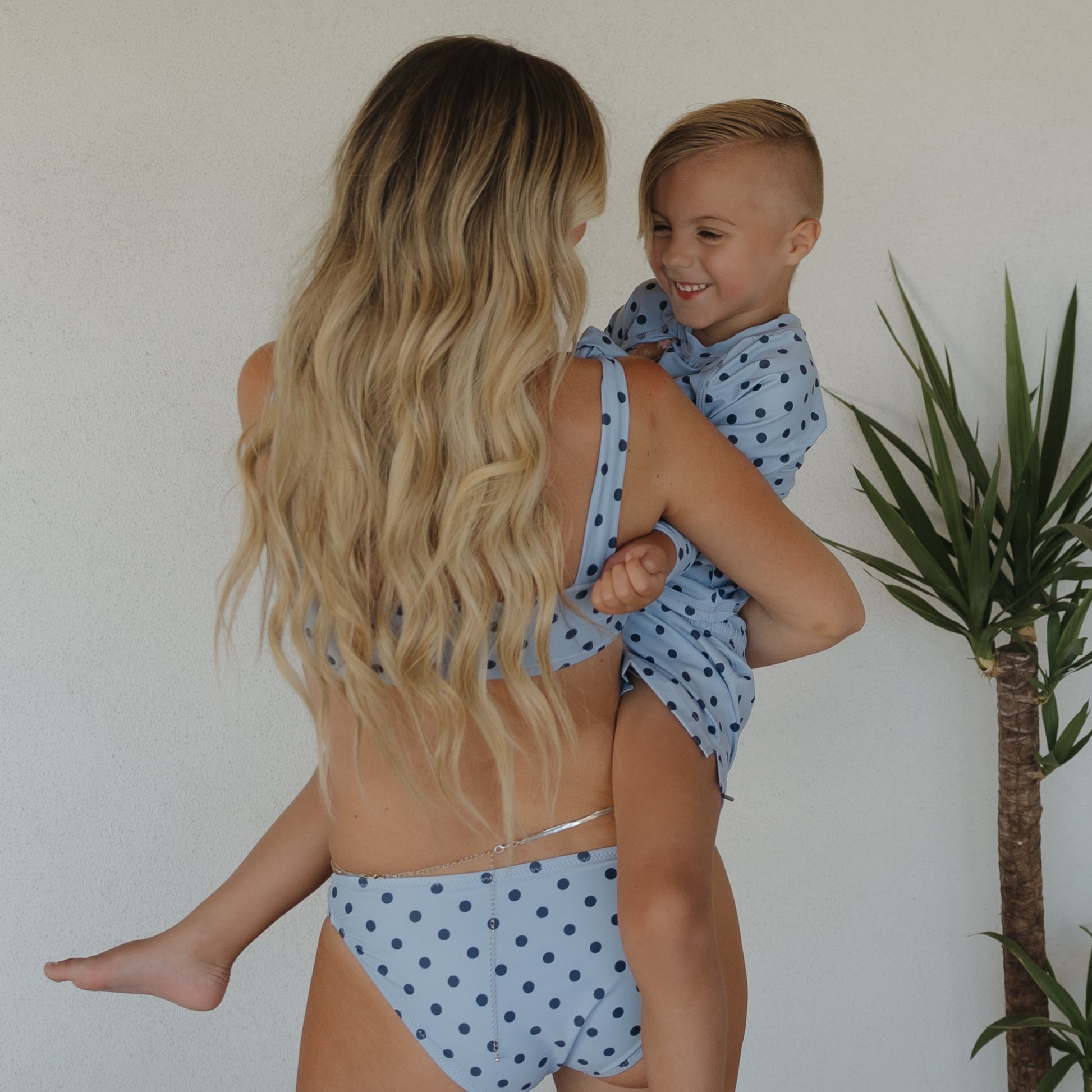 A woman with long blonde hair wears the forever french baby Women's Two Piece Swimsuit | Sully, matching her smiling child in family swimwear, as they stand by a white wall with a potted plant nearby.