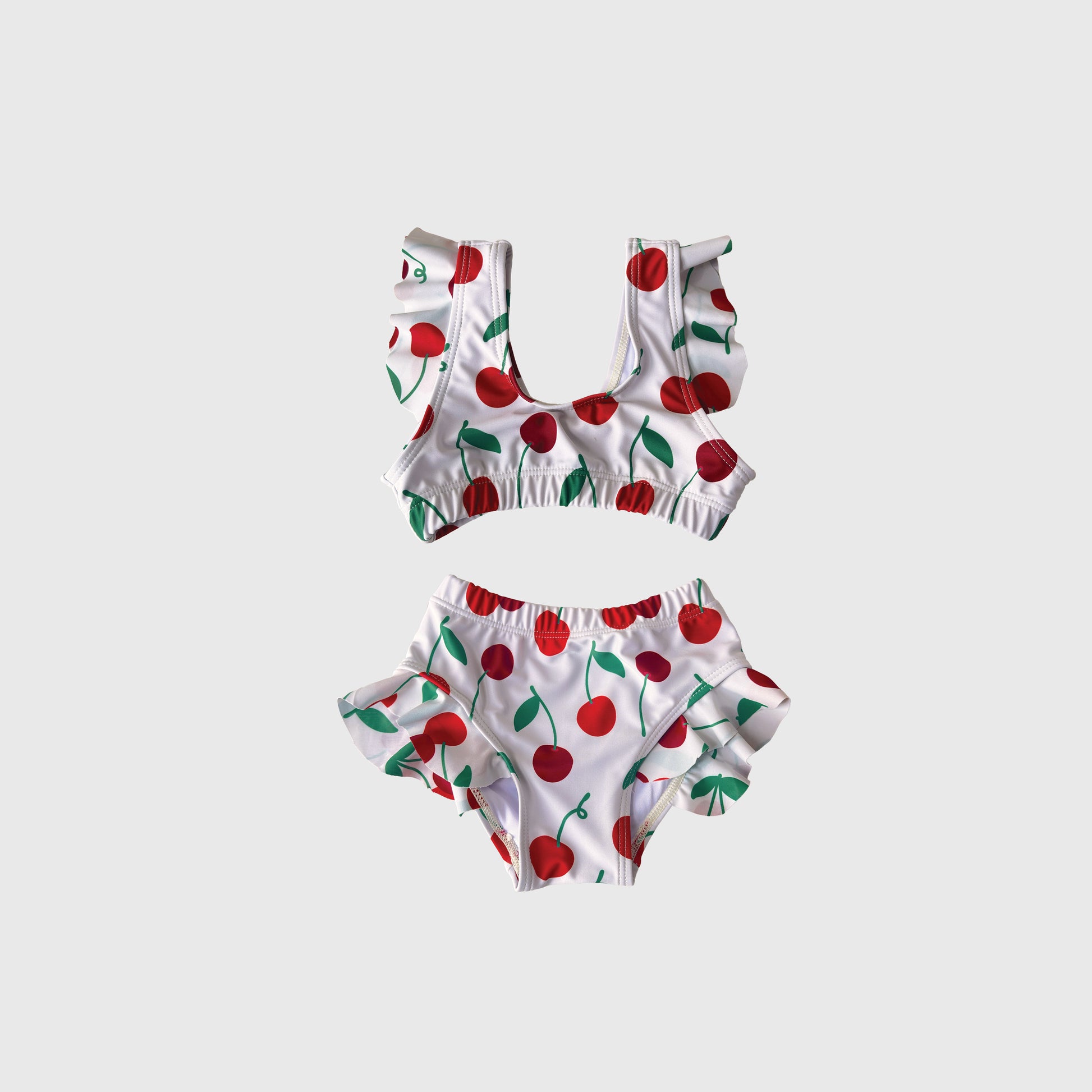 The Girl's Two Piece Swimsuit | Cherry by forever french baby features a white ruffled crop top and high-waisted bottoms with a red cherry and green leaf print, set against a light gray background.