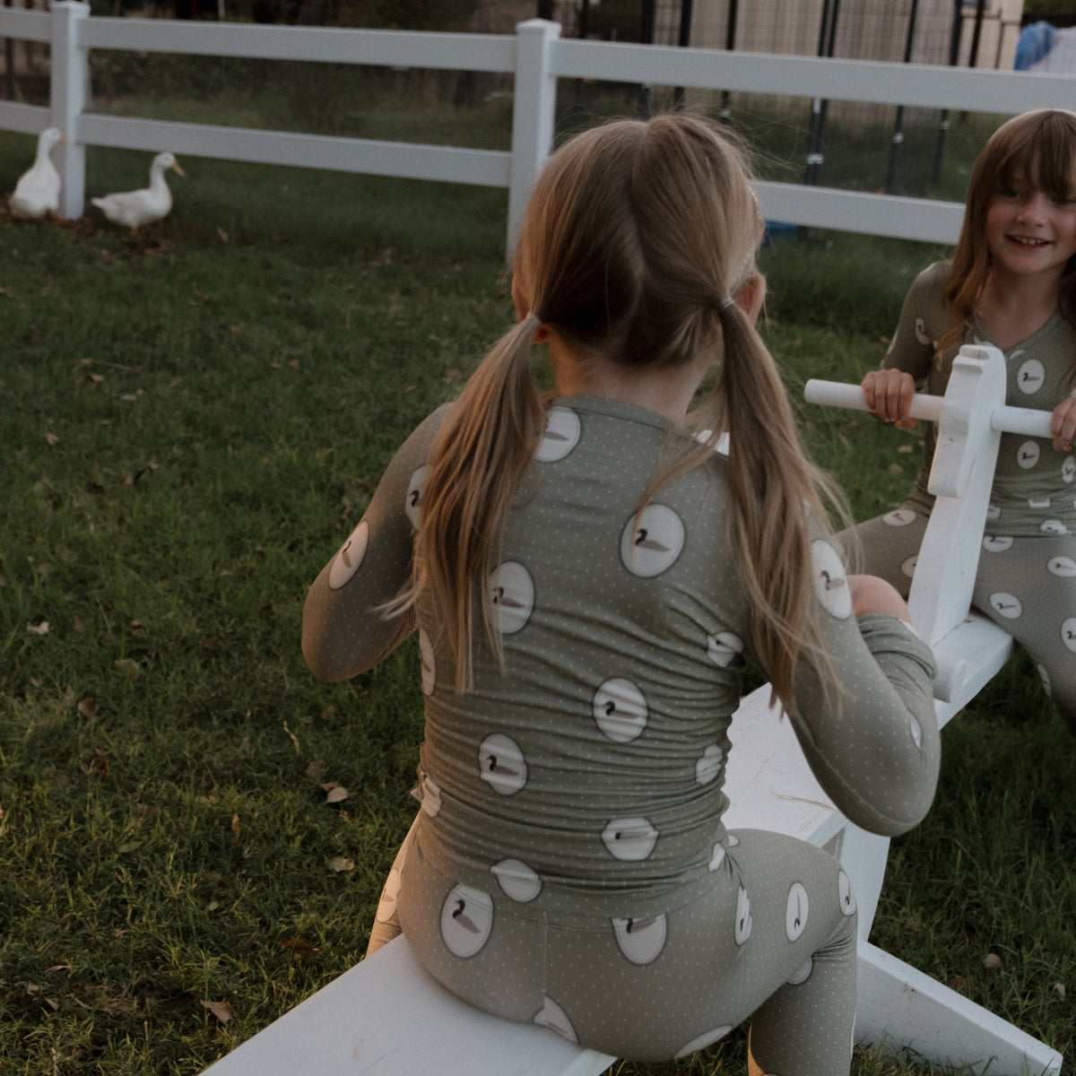 Two young children wearing matching Bamboo Two Piece Pajamas in the Mallard Frames print by forever french baby play on a white seesaw outdoors. Green grass, a white fence, and two white ducks are visible in the background—the perfect setting for cozy, hypoallergenic sleepwear crafted from kids’ bamboo clothing.