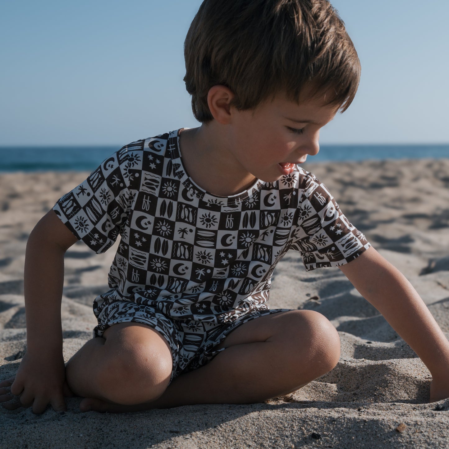 A young boy sits on the sandy beach, playing as he wears the forever french baby Bamboo Short Set | Summer Check, a breathable black and white patterned outfit. The sea and sky stretch out in the background.