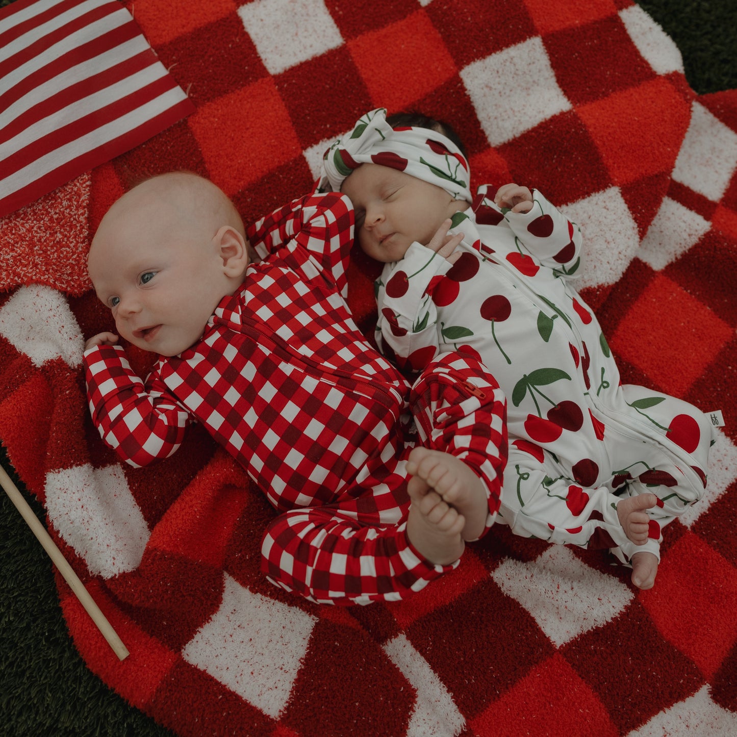 Two babies lie on a red and white checkered blanket—one in a red checkered onesie, the other cozy in forever french baby's Bamboo Zip Pajamas | Cherry, featuring cherry prints and a matching hat. An American flag is placed beside them.