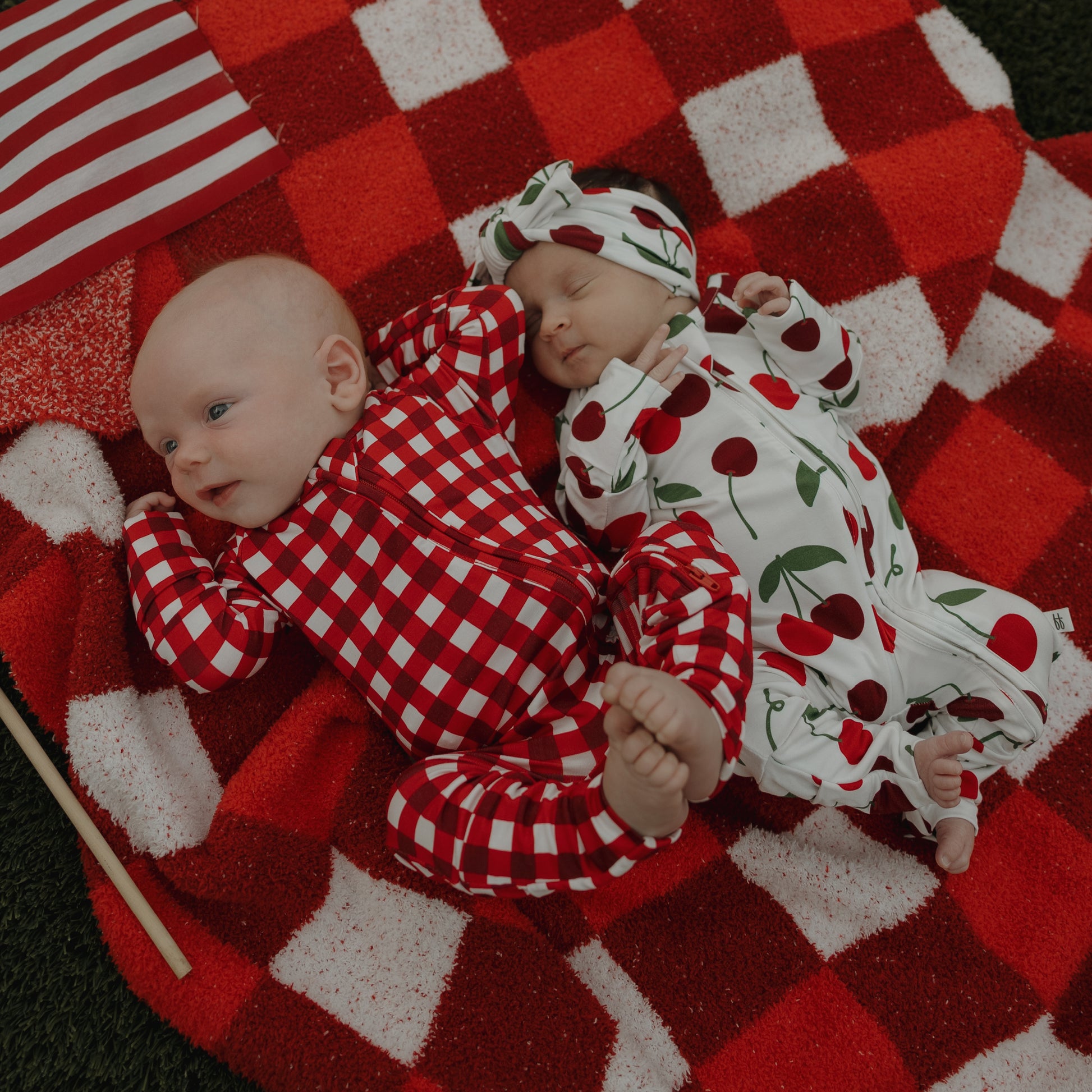Two babies lie on a red and white checkered blanket—one in a red checkered onesie, the other cozy in forever french baby's Bamboo Zip Pajamas | Cherry, featuring cherry prints and a matching hat. An American flag is placed beside them.