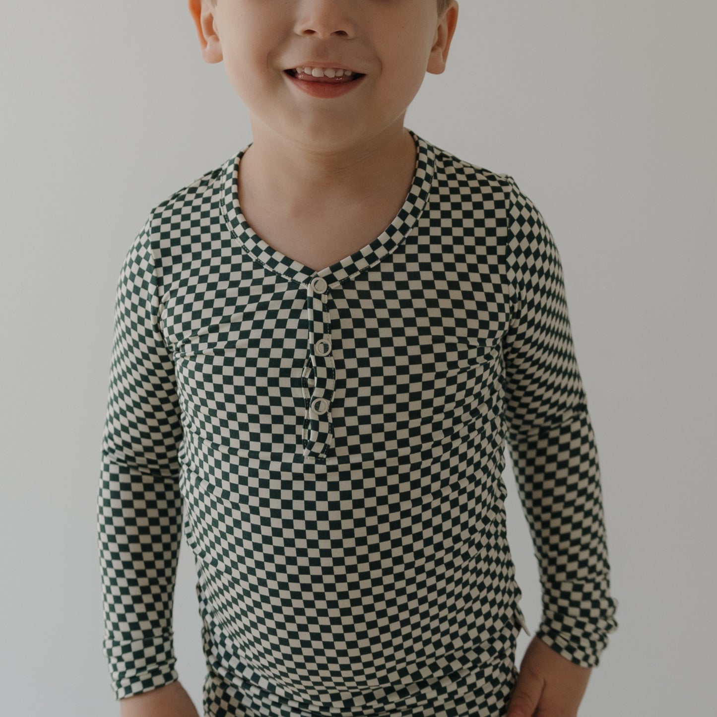 A young child smiles in forever french baby's Bamboo Two Piece Pajamas | Lucky Micro Check, featuring a black-and-white checkered pattern and hypo-allergenic fabric, against a plain white background.