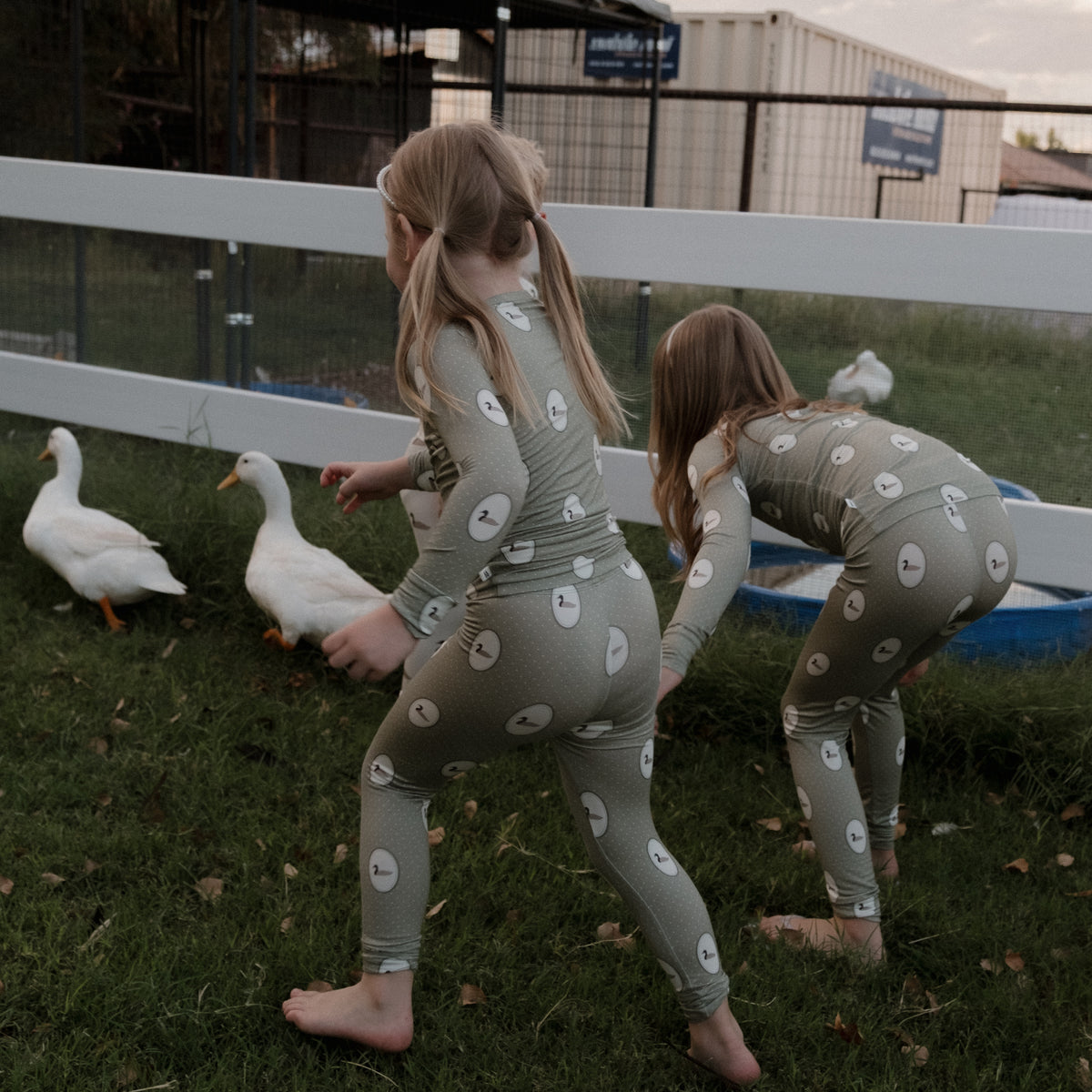 Wearing forever french baby Bamboo Two Piece Pajamas in the Mallard Frames print, two young girls crouch barefoot on the grass, approaching three white ducks by a fenced area beneath an overcast sky.
