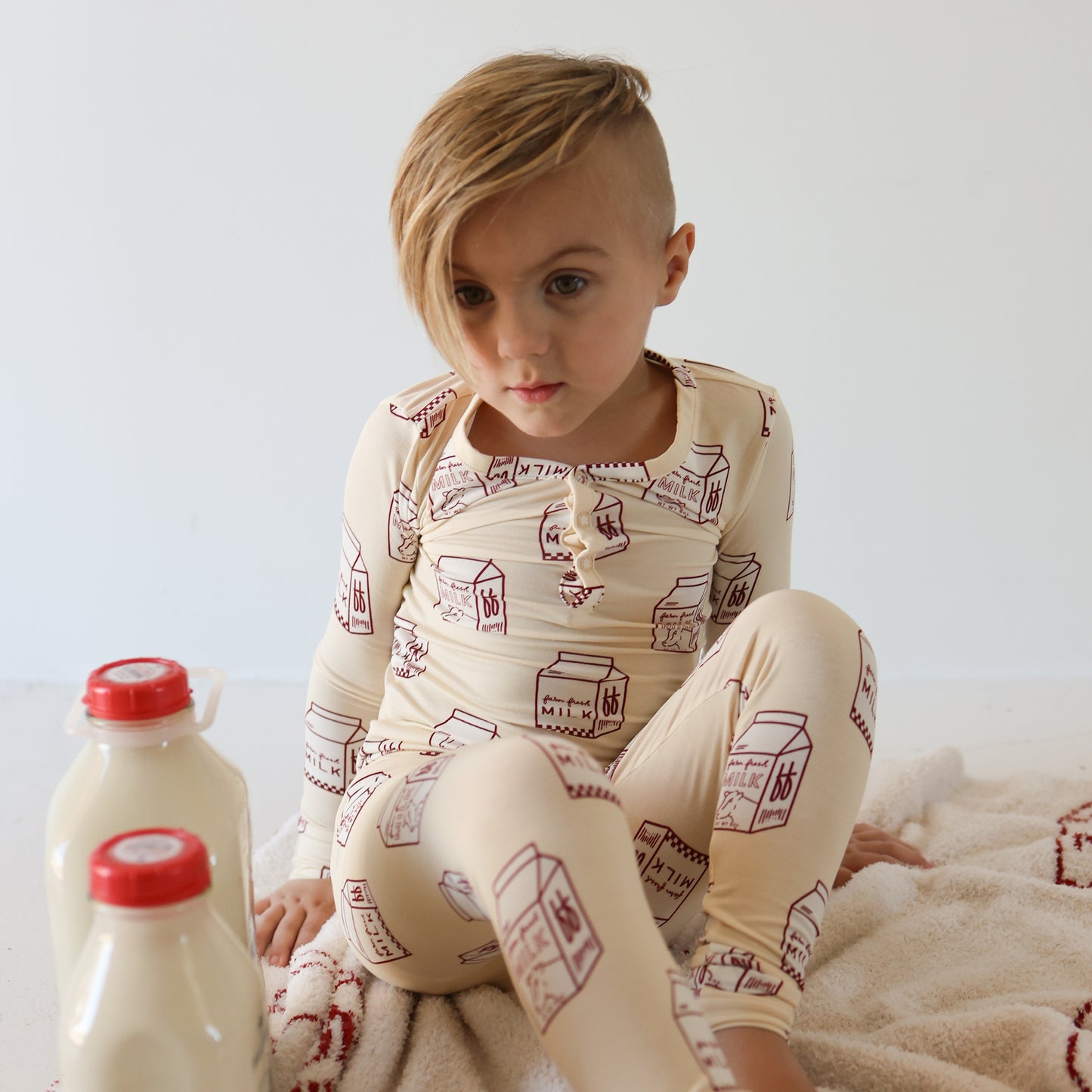 A young child with an asymmetrical haircut sits on a soft surface wearing forever french baby’s Bamboo Two Piece Pajamas | Milk, decorated with milk carton prints. Two bottles of milk with red caps are nearby on the floor.