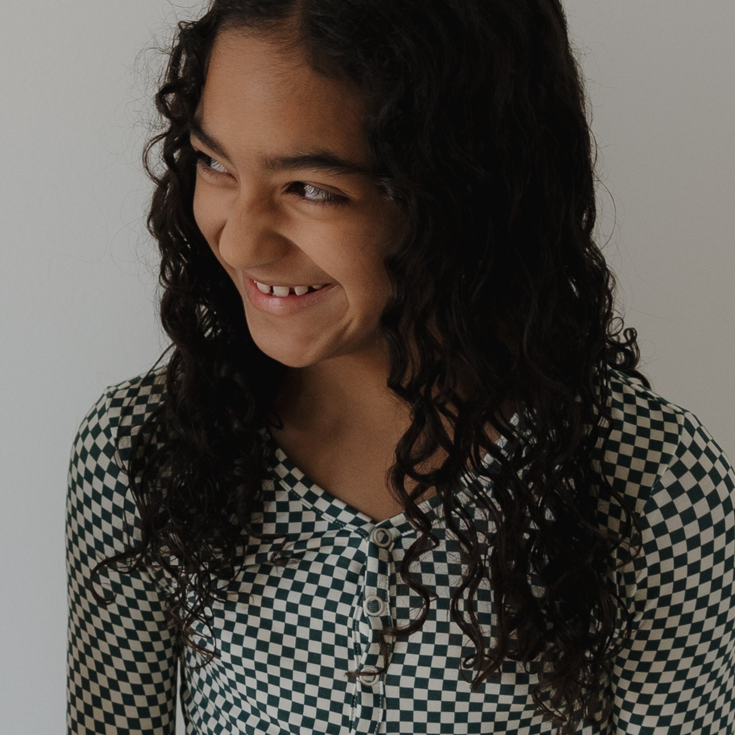 A young person with long, curly dark hair smiles and looks to the side, wearing forever french baby’s Bamboo Two Piece Pajamas in Lucky Micro Check against a plain, light background.