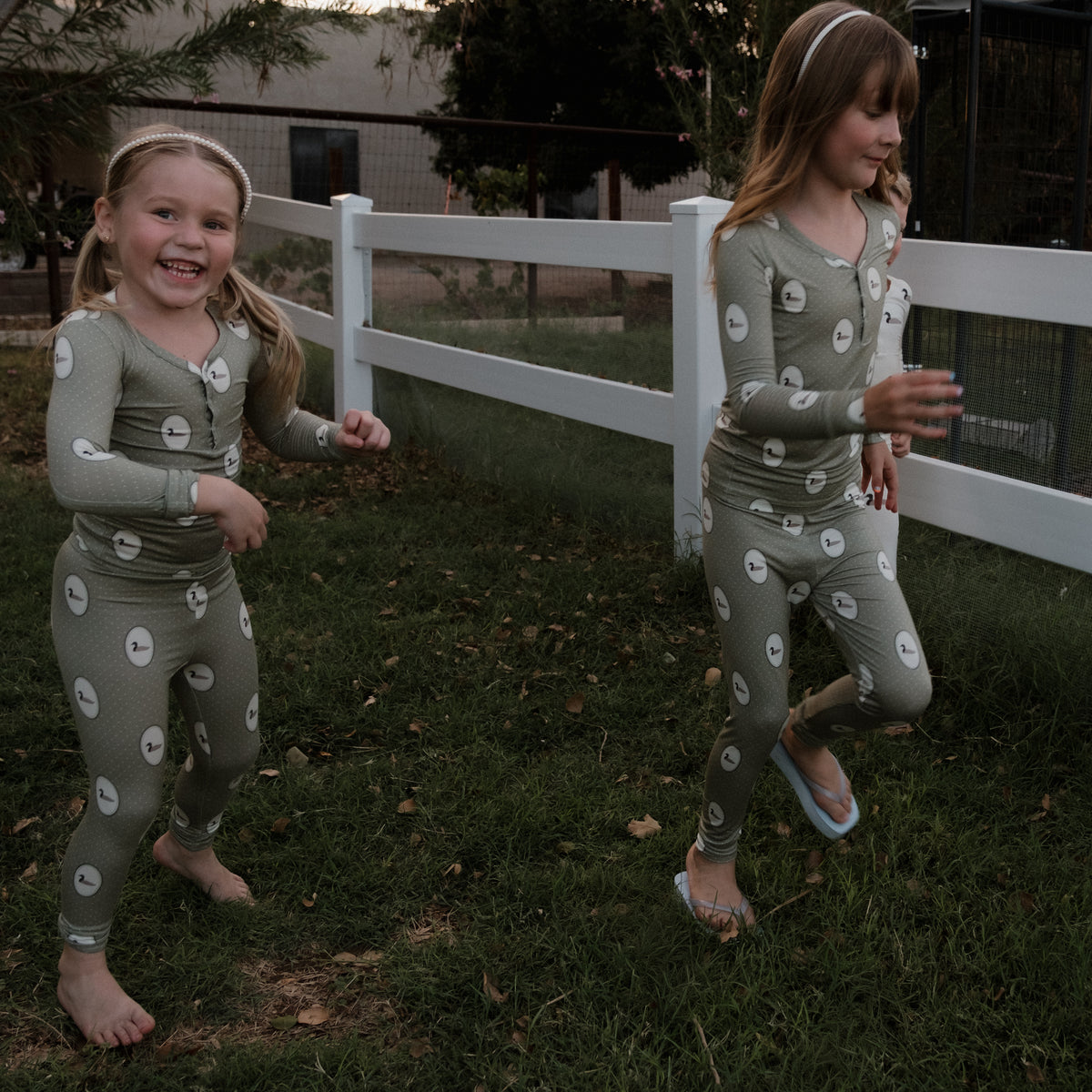 Two young girls wearing matching forever french baby Bamboo Two Piece Pajamas in the Mallard Frames print—featuring a green bamboo fabric with playful white circle patterns—are running and playing barefoot on the grass near a white backyard fence. These pajamas are stylish, gentle on sensitive skin, and hypoallergenic, making them the perfect choice for kids’ sleepwear.