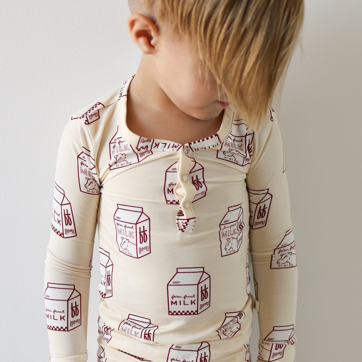 A young child with blond hair stands against a white wall, wearing forever french baby's Bamboo Two Piece Pajamas | Milk, cream-colored, breathable kids sleepwear decorated with red milk carton illustrations.