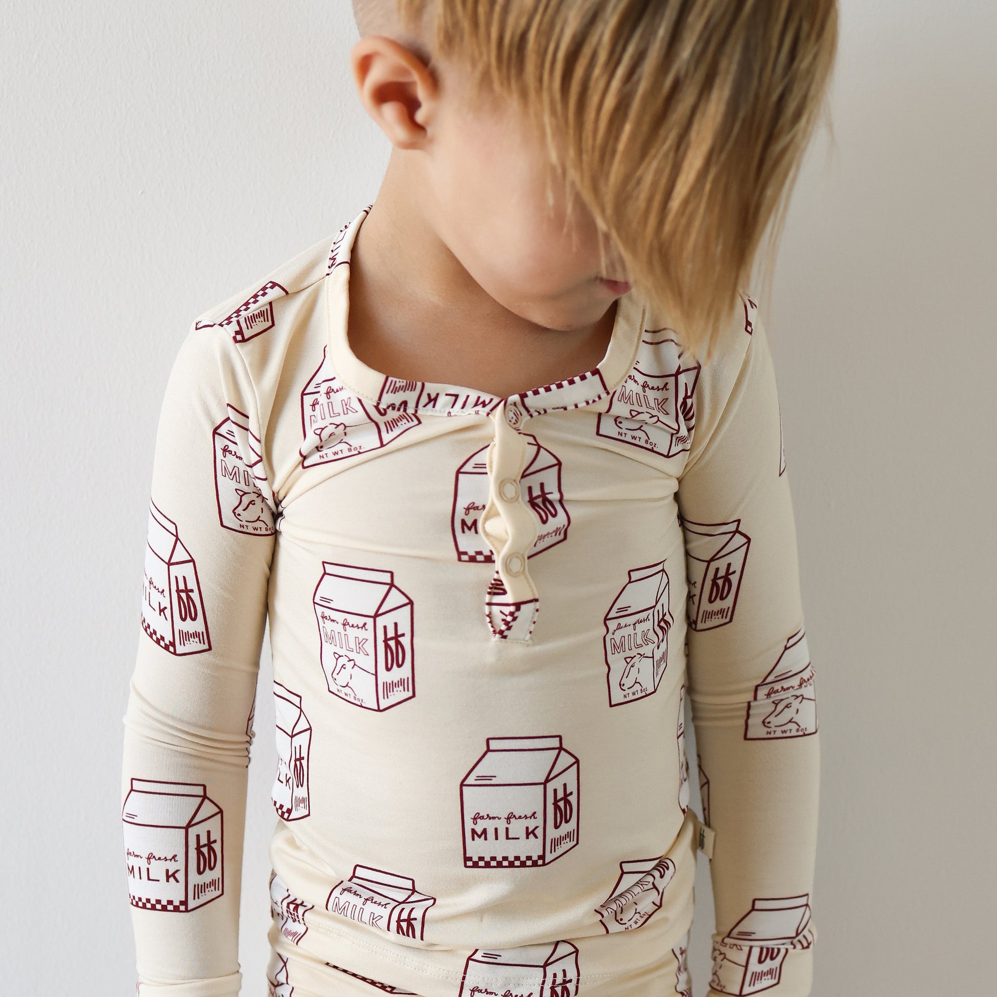 A young child with blond hair stands against a white wall, wearing forever french baby's Bamboo Two Piece Pajamas | Milk, cream-colored, breathable kids sleepwear decorated with red milk carton illustrations.
